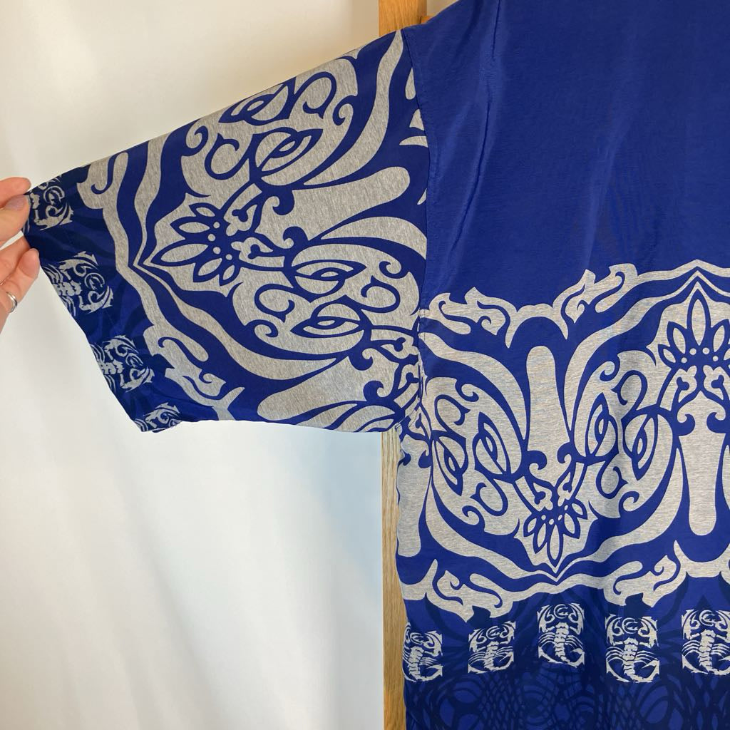 A royal blue kaftan-style tunic with wide sleeves, featuring an ornate white and silver floral and scroll pattern, held out by a hand on the left side against a white background.