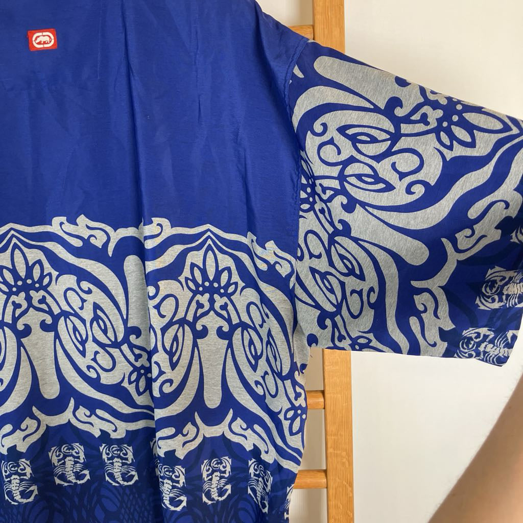 A royal blue kaftan-style tunic with a wide silver-gray ornamental border and small scorpion motifs near the hem, shown hanging on a wooden hanger against a light wall.