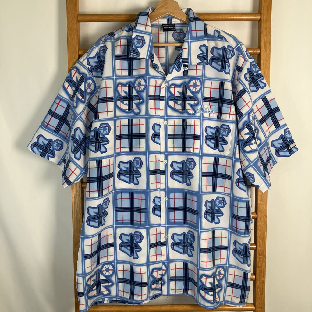 A short-sleeve button-up shirt with a blue, white, and red plaid pattern hanging on a wooden hanger against a light-colored wall.