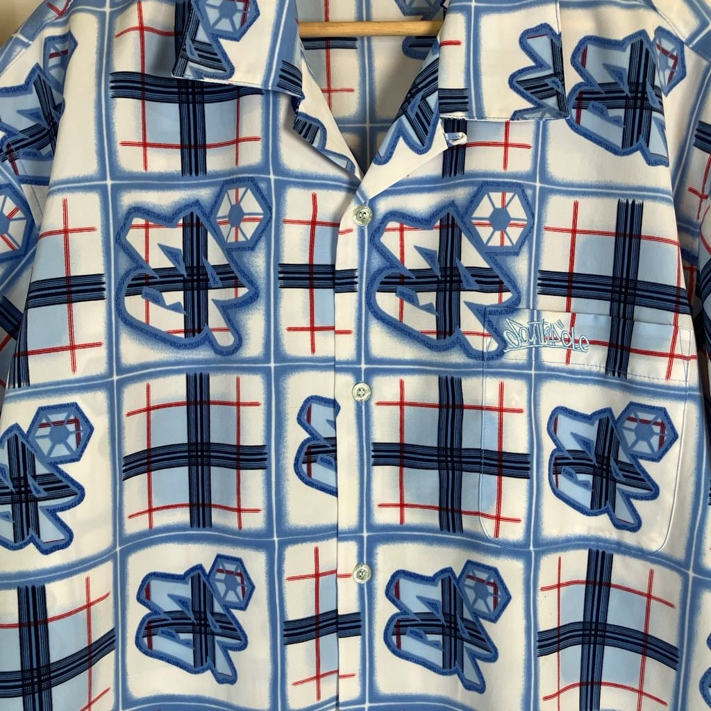 A close-up of a button-front shirt with a white base and a repeating pattern of blue and navy plaid squares, red grid lines, and blue geometric star-like motifs. A wooden hanger is visible at the collar, and a small white script logo appears near the right chest area.