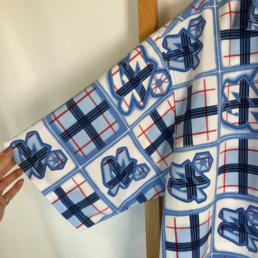 A close-up of a white kimono-style garment with a blue and red plaid grid pattern and stylized blue motifs, held by a hand against a plain indoor background.