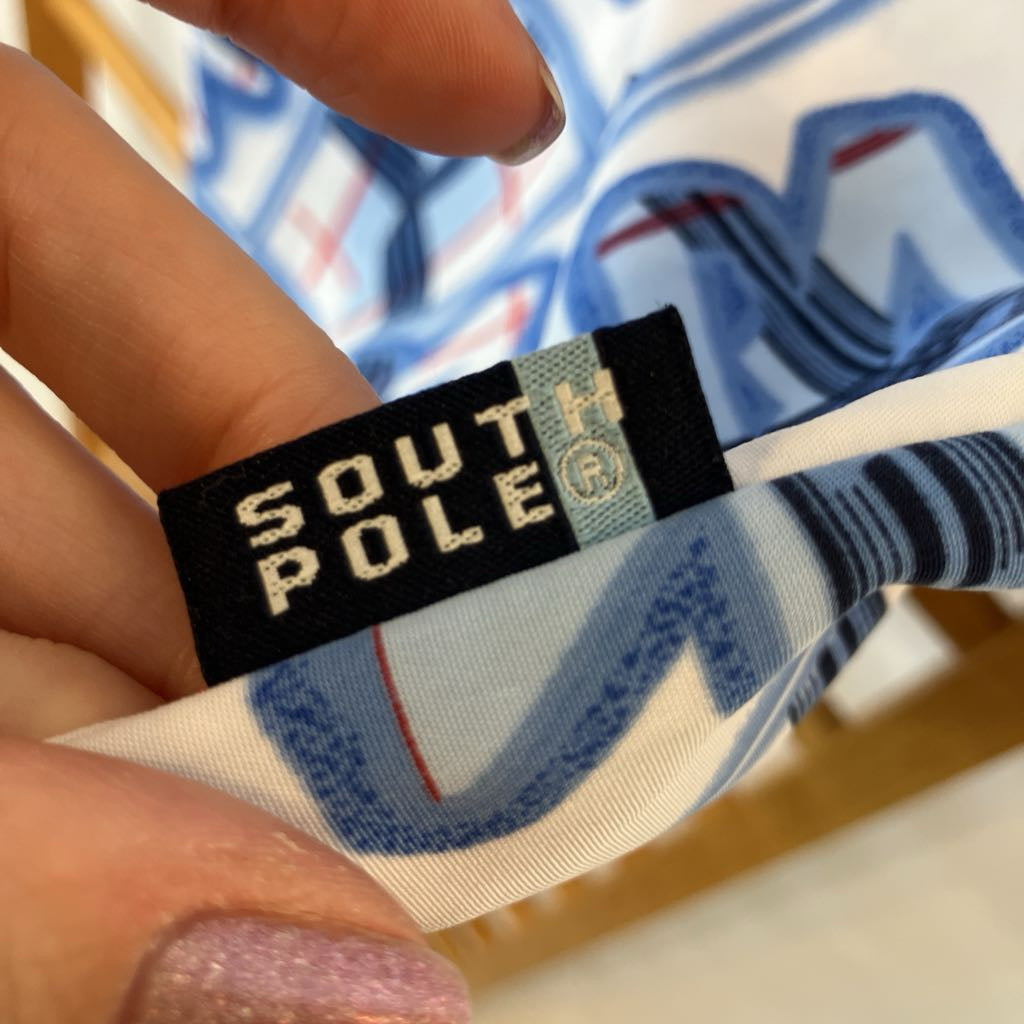 A close-up of a hand holding a white fabric item with a blue graphic print, showing a black and light blue South Pole brand tag sewn onto the inside.