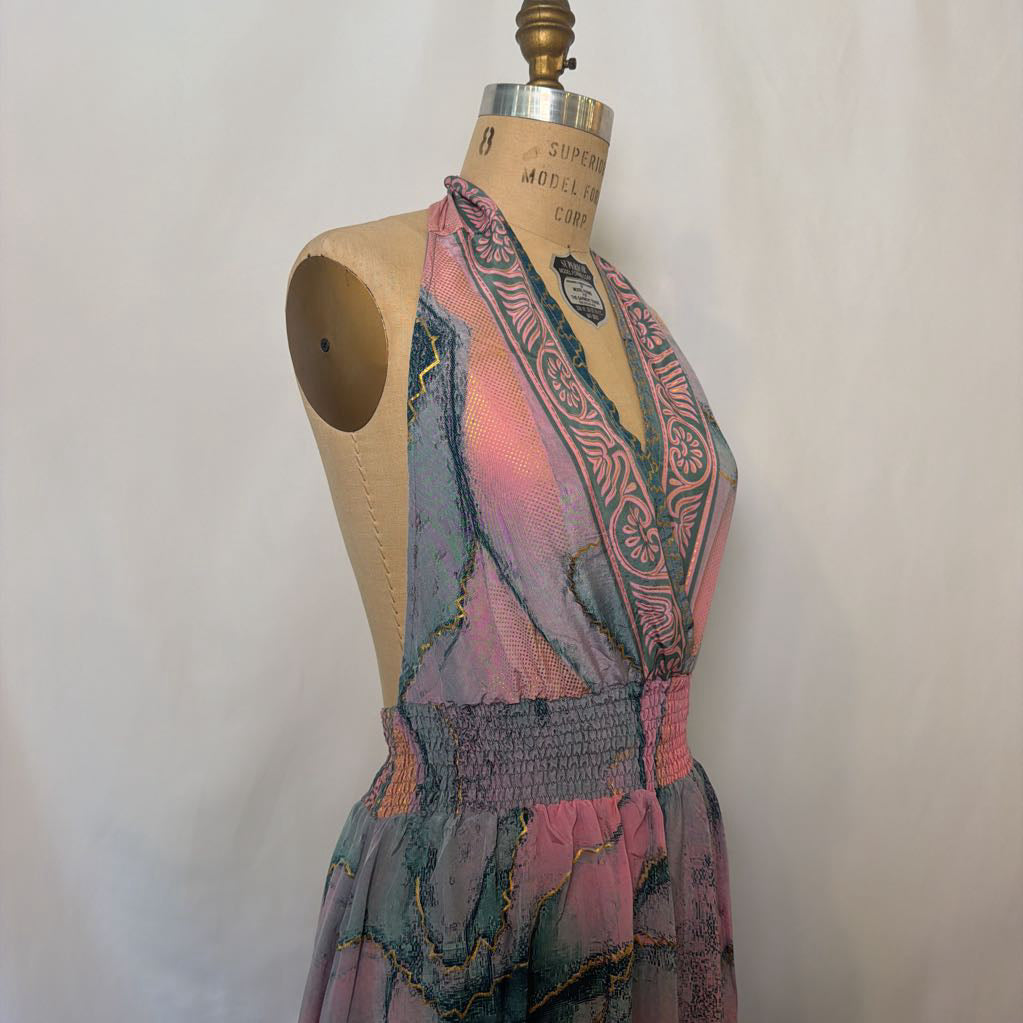 A sleeveless halter-neck dress displayed on a wooden dress form mannequin. The dress features a vibrant, painterly abstract pattern in shades of pink, teal, blue, and purple with gold accents, and has a smocked elastic waist.