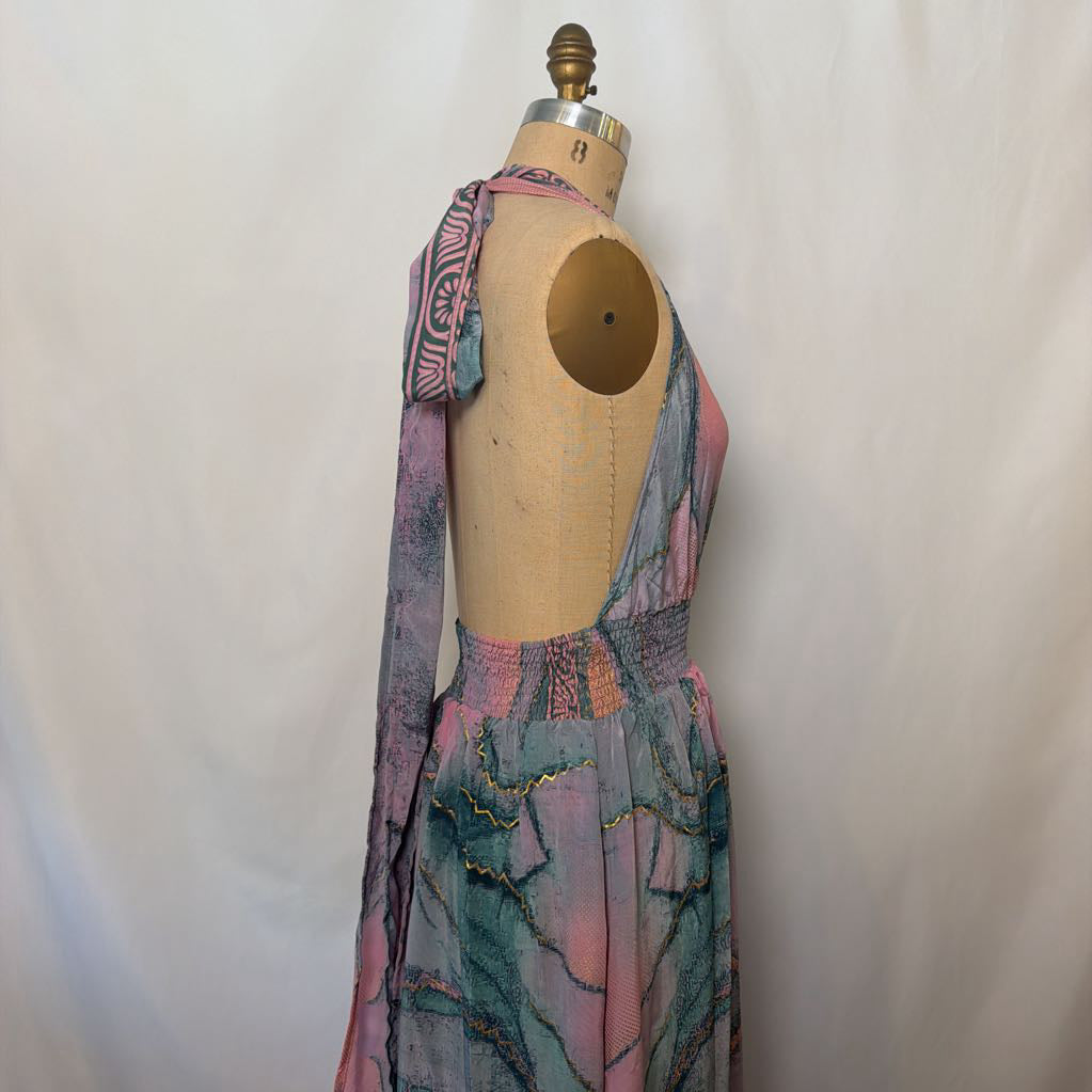 A sleeveless halter dress displayed on a vintage wooden dress form mannequin against a white fabric backdrop. The dress features a multicolor abstract print with pink, teal, and purple tones, and gold sequin detailing along the waist and skirt.