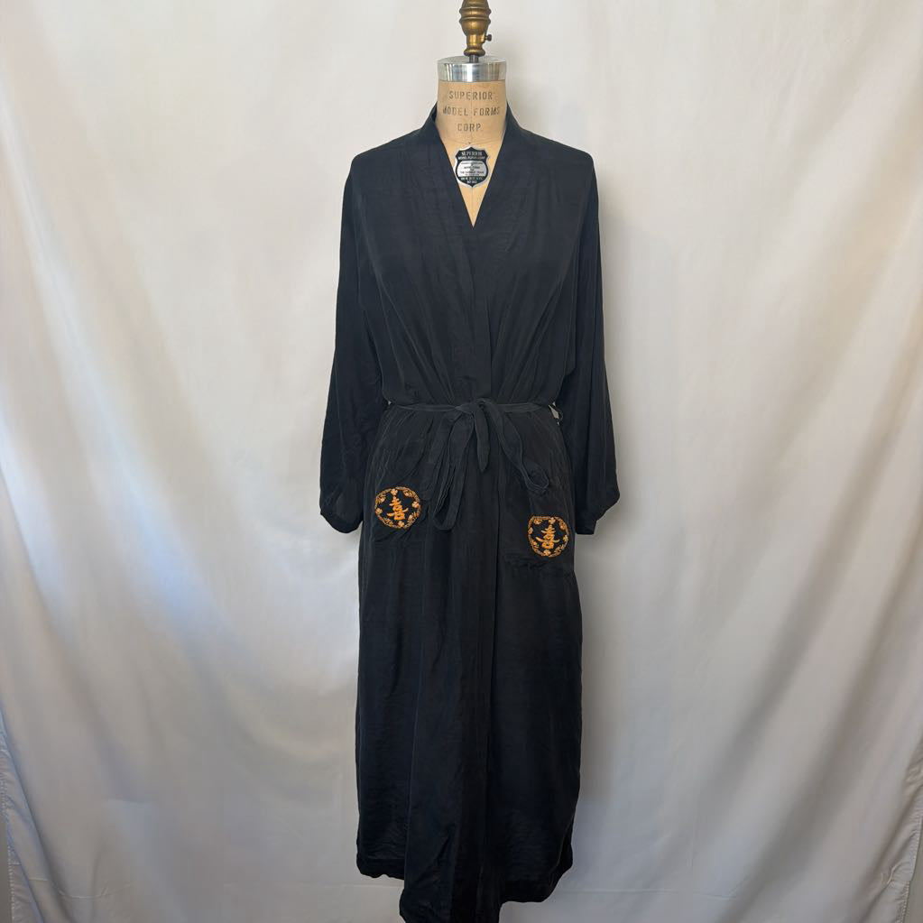 A black long robe displayed on a dress form against a white fabric backdrop. The robe has a tie belt and two circular embroidered patches near the lower front.