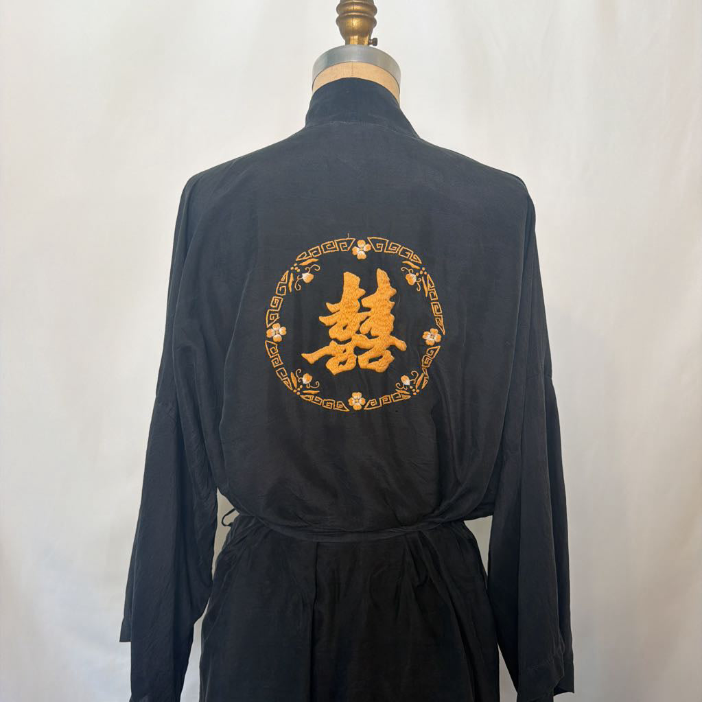 A black long-sleeve robe or kimono-style garment displayed on a dress form from the back, featuring a large gold embroidered Chinese double happiness symbol inside a decorative circular border.