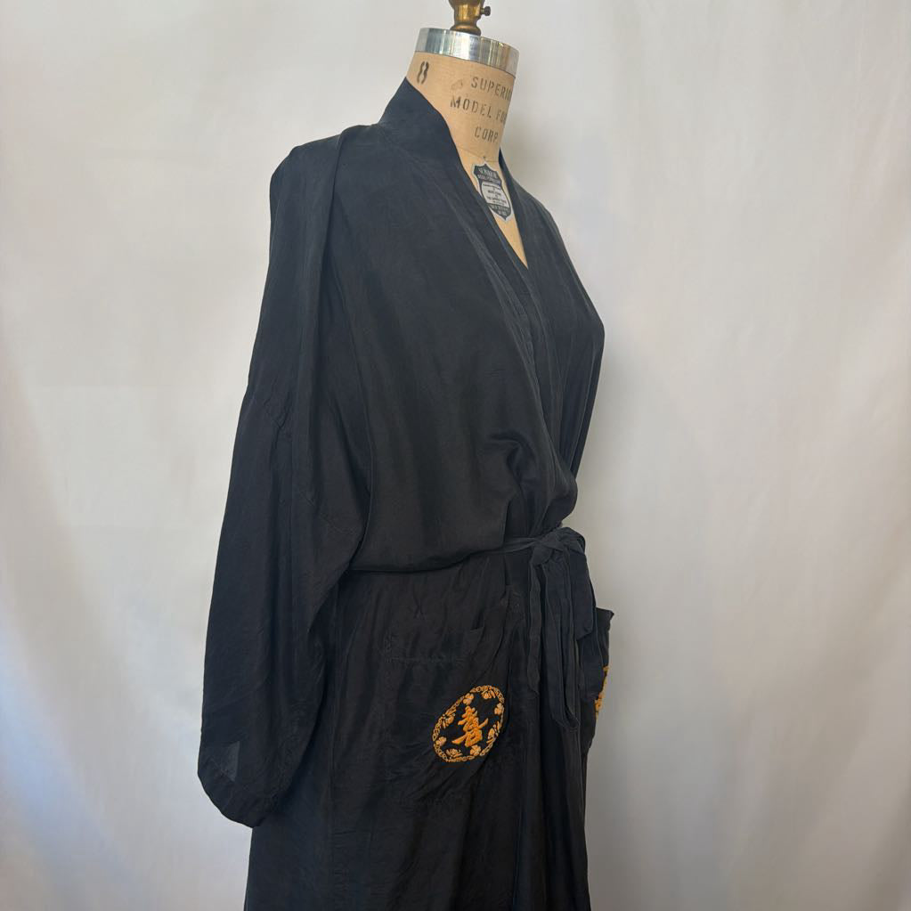 A black kimono-style robe displayed on a dress form mannequin against a light gray fabric backdrop. The robe has a tie belt and a circular gold embroidered patch on the lower front.