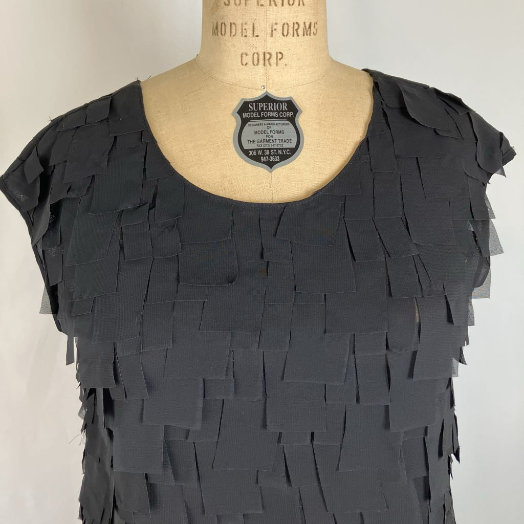 A black fringed top displayed on a beige dress form mannequin against a plain light background. The top features a scoop neckline and cascading rectangular fringe layers across the front.