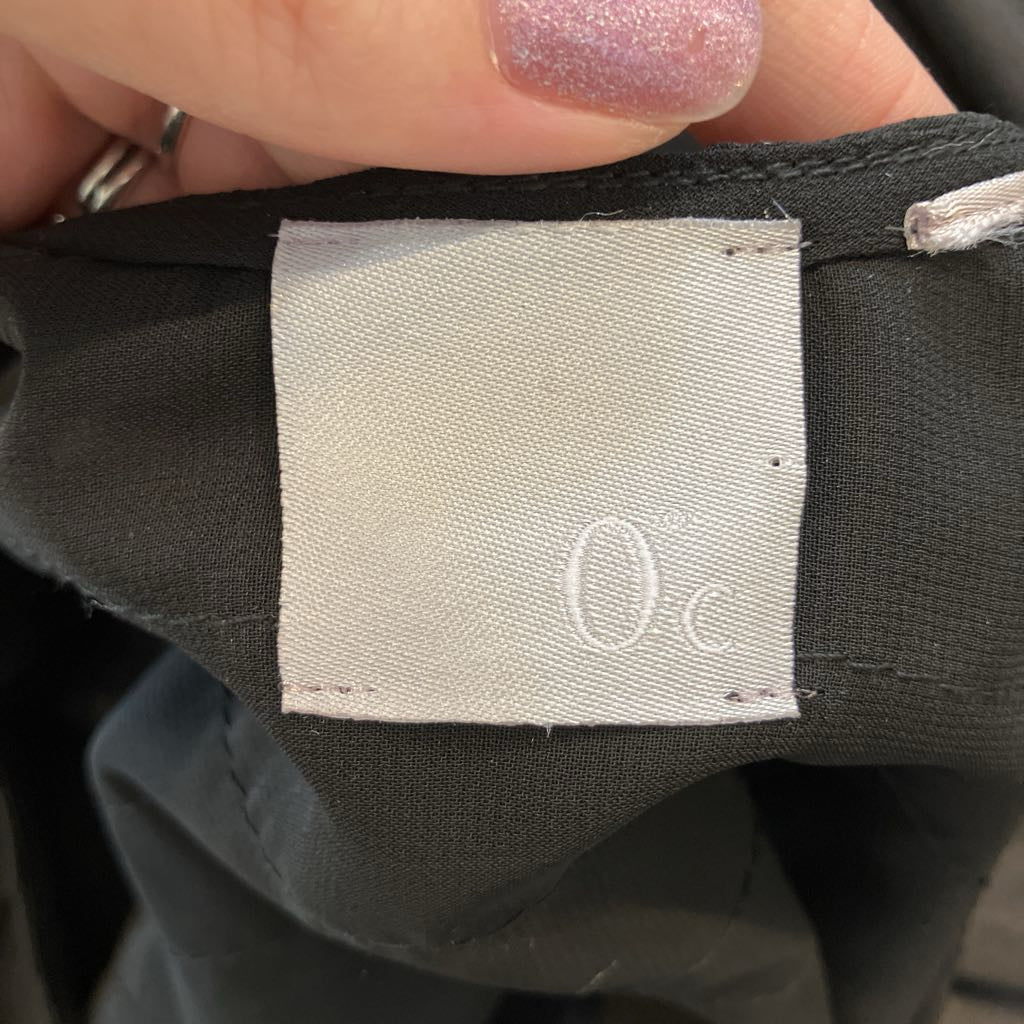 Close-up of a garment interior with a white fabric label reading 'Oc' sewn into dark charcoal fabric, held by a hand with glittery pink nail polish.
