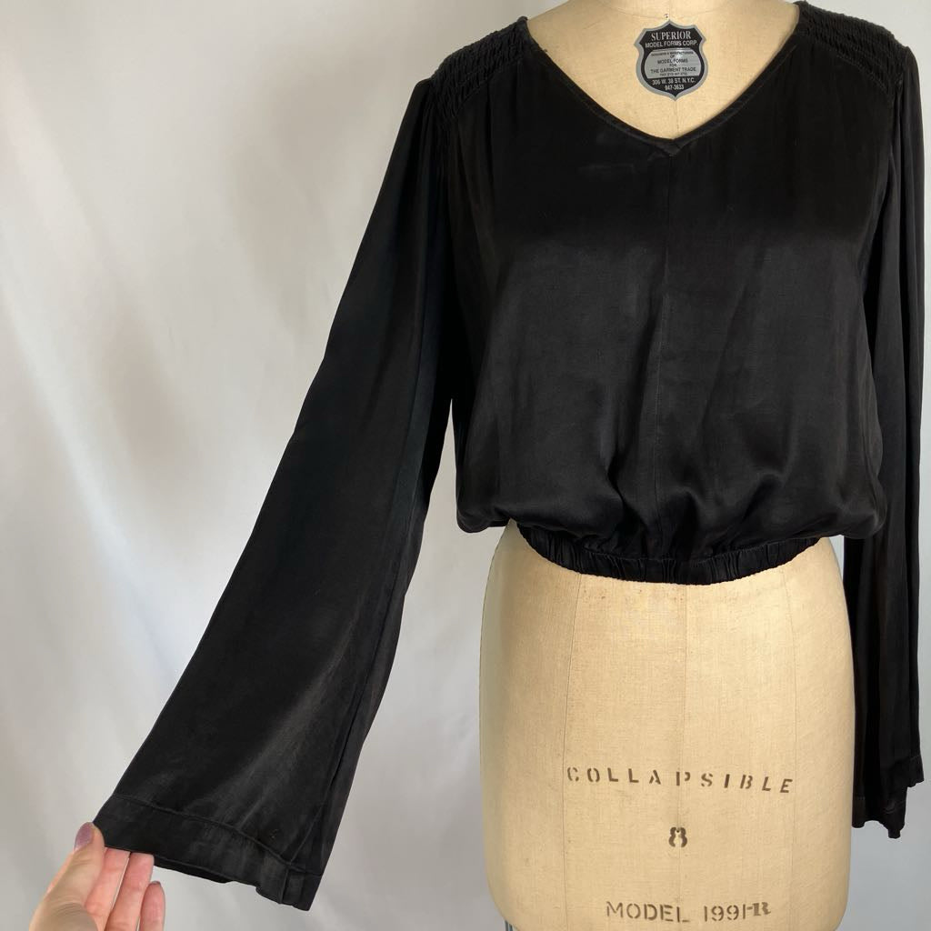 A black long-sleeve blouse displayed on a beige dress form mannequin against a white fabric backdrop. The blouse features a V-neckline and a gathered waist with a smocked or elasticized finish.