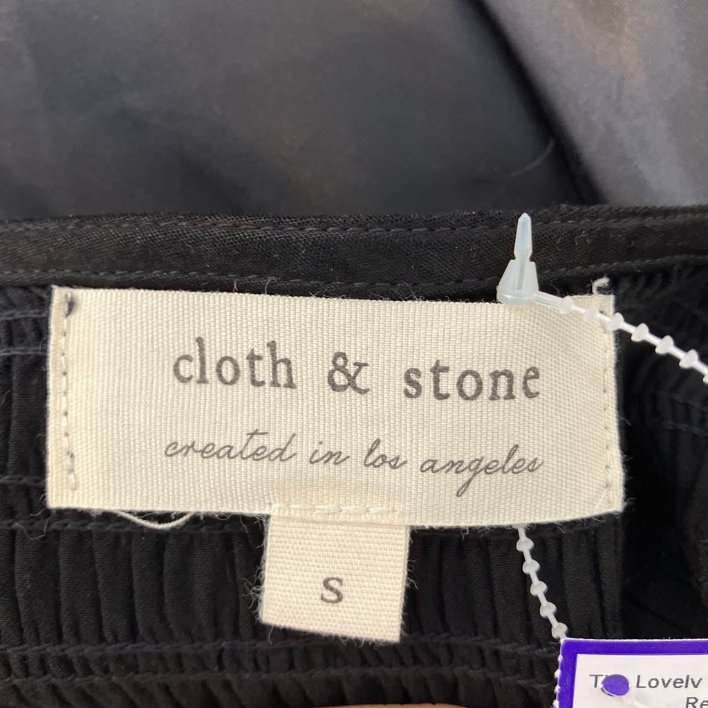 Close-up of a black ribbed garment label reading 'cloth & stone created in los angeles' with a size 'S' tag below, photographed against dark fabric.