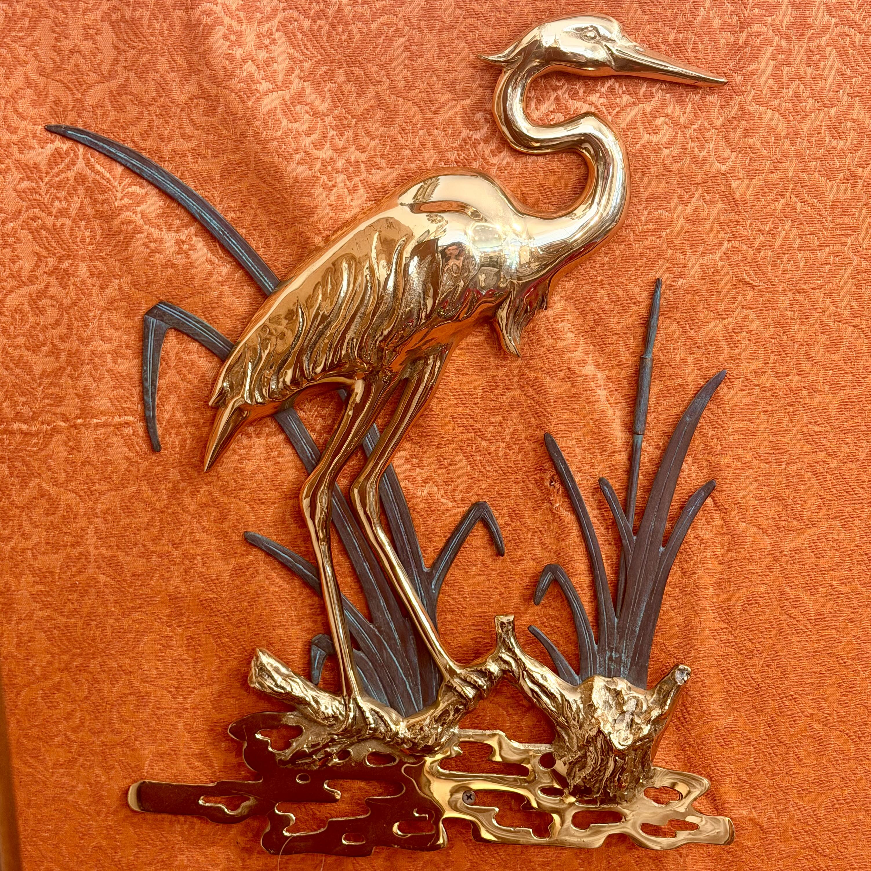 A decorative wall sculpture of a heron standing among reeds on a textured orange background.
