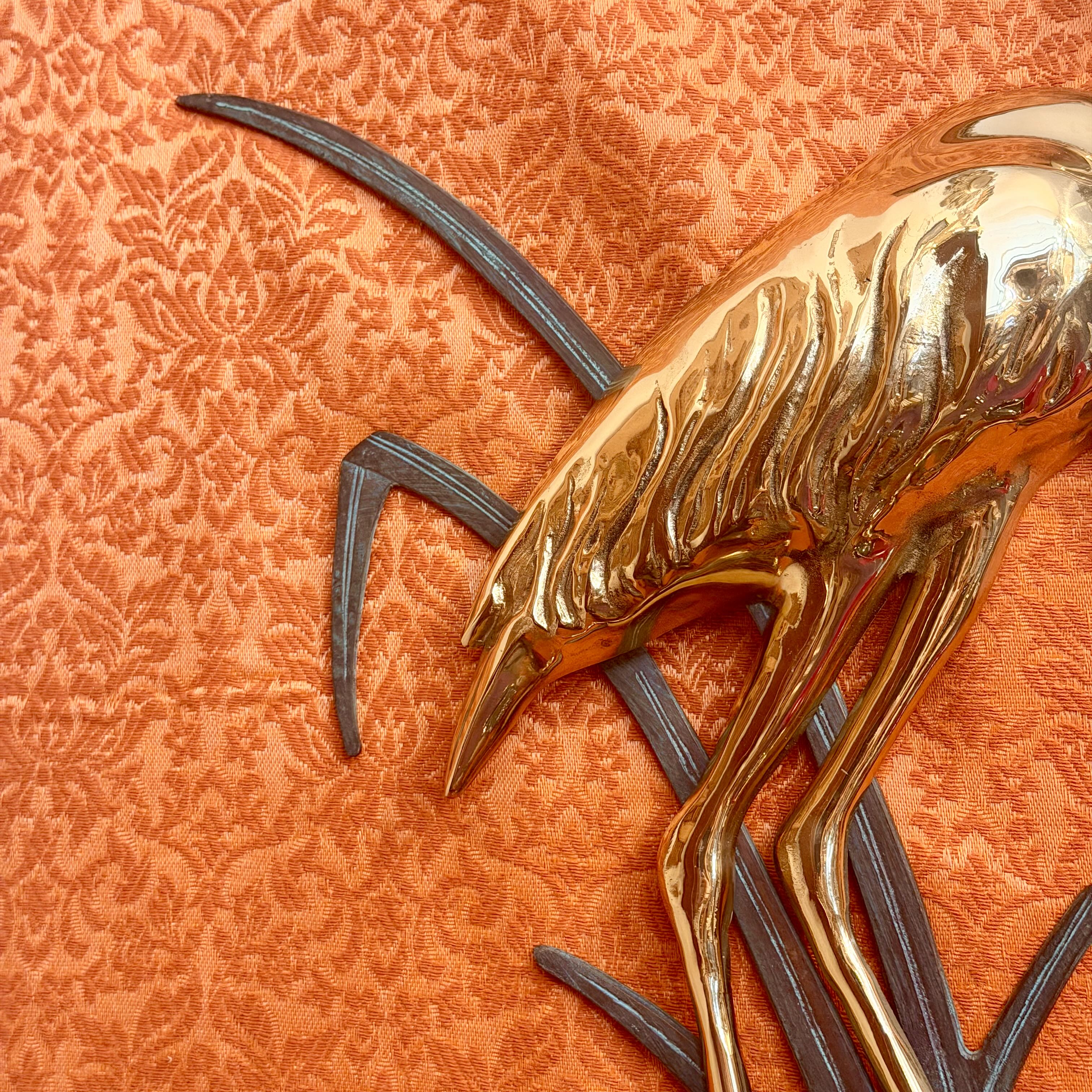 A close-up of a polished gold-toned decorative bird sculpture with dark gray branch-like elements, set against a textured orange damask fabric background.