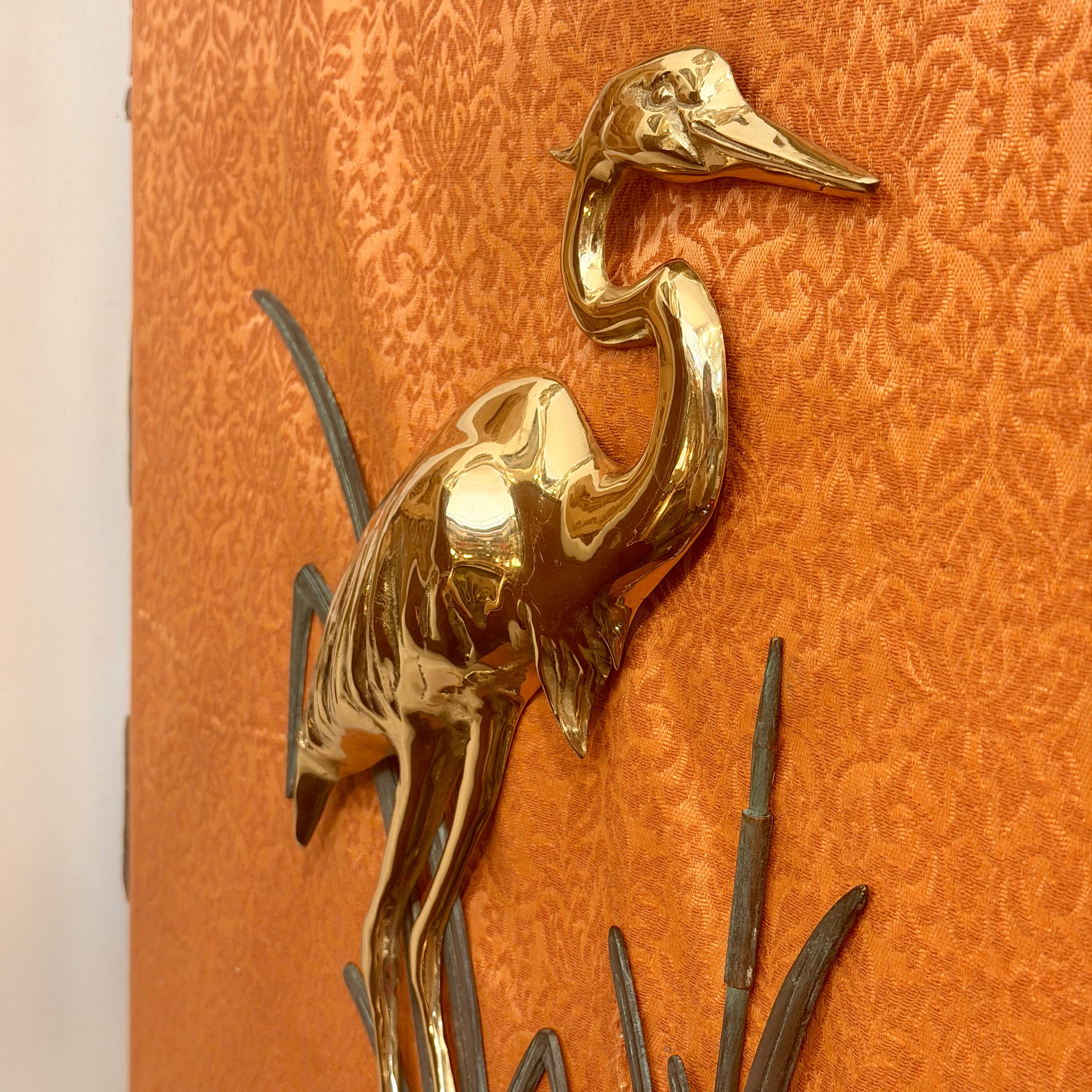 A polished gold-toned metal heron sculpture mounted on a textured orange damask wallpaper background.