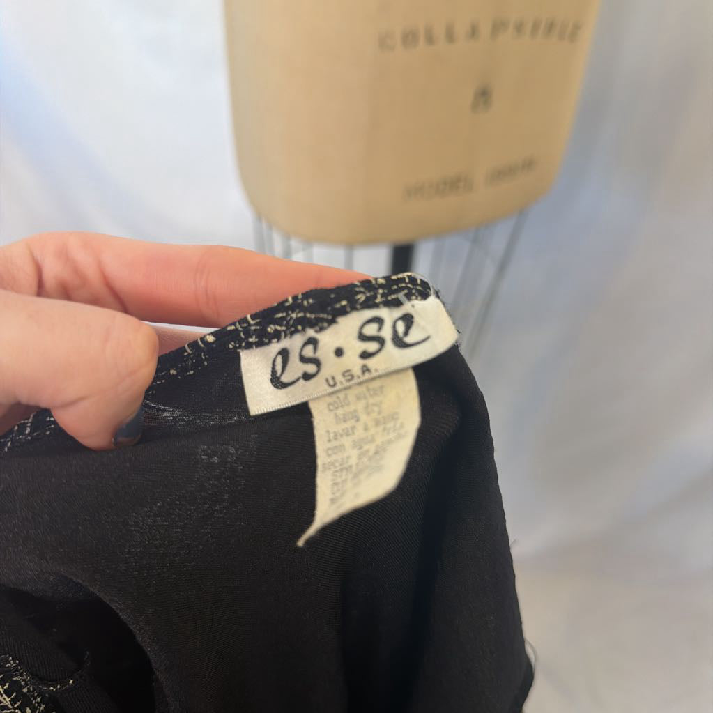 A close-up of a person's hand holding the collar of a black garment, showing the brand label 'es.se USA' and size 'B'.