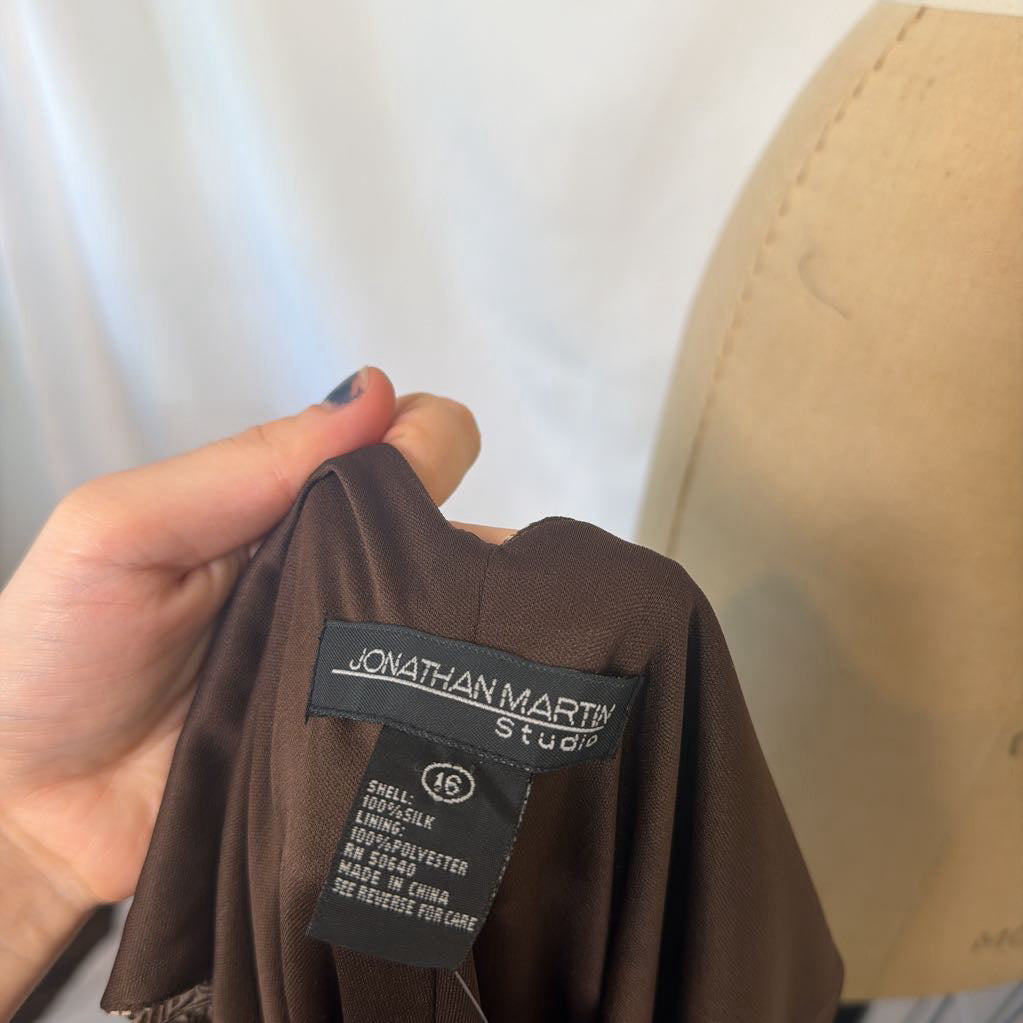 A close-up of a person's hand holding a brown garment, showing the interior neck label which reads 'JONATHAN MARTIN Studio'.