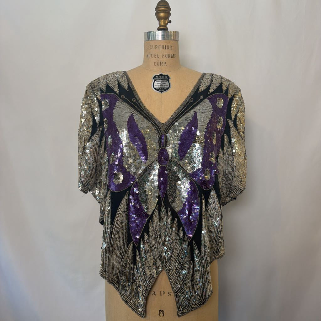 A vintage sequined blouse displayed on a wooden dress form. The top features a silver and black beaded pattern with large purple sequined butterfly wings on the front.