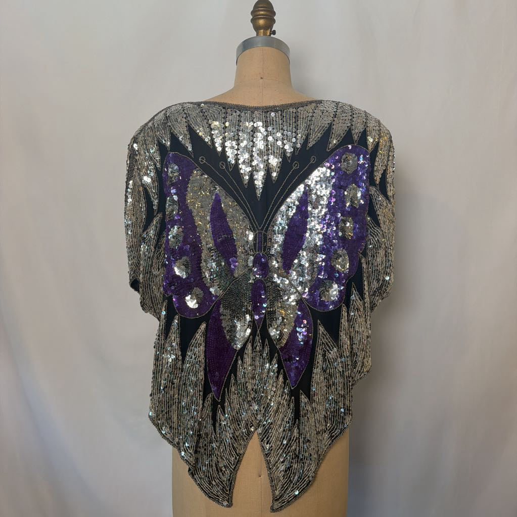 A sequined dance or performance top displayed on a dress form mannequin. The back of the top features a large butterfly design embellished with silver and purple sequins on a black base, set against a neutral light gray fabric backdrop.