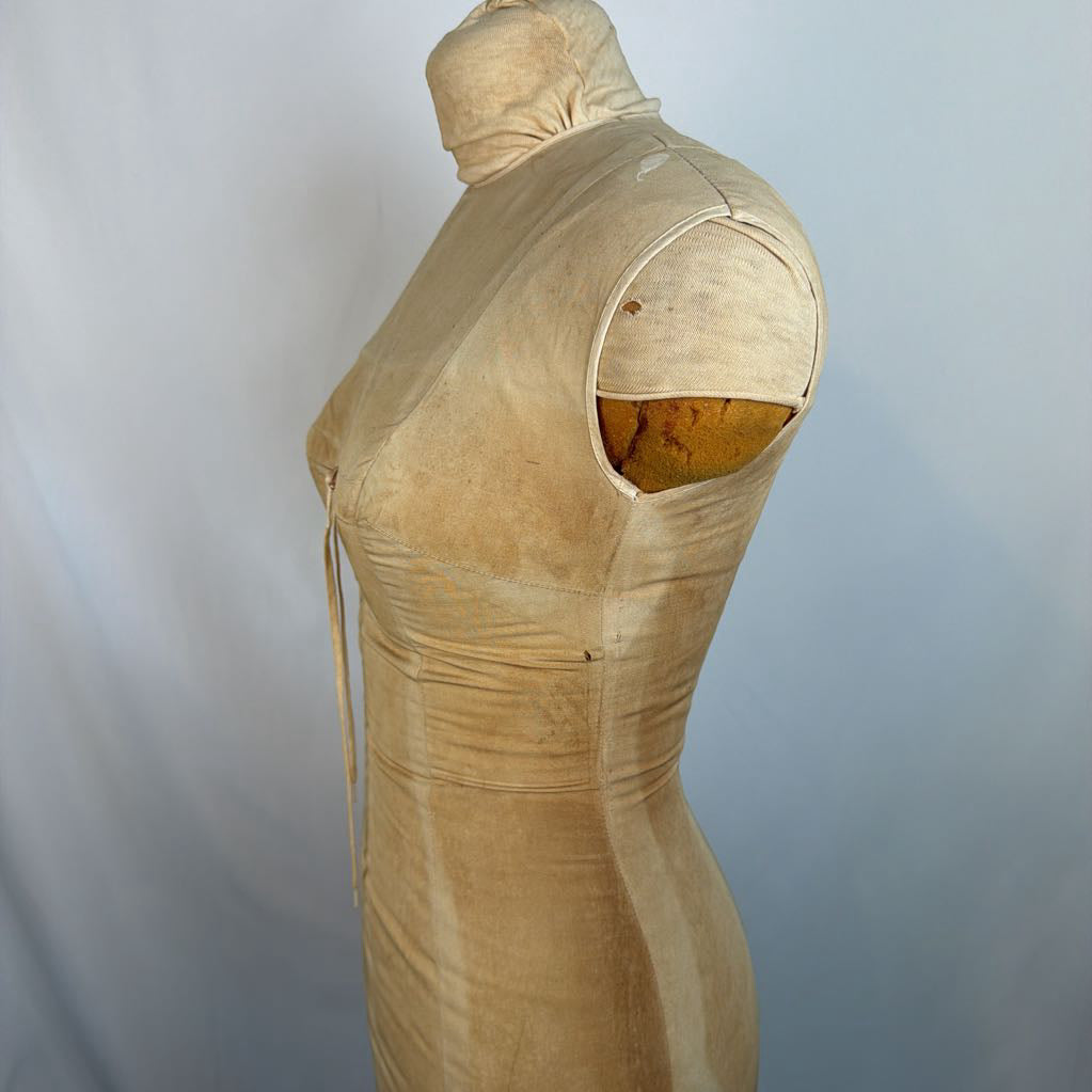 A beige sleeveless dress or costume displayed on a mannequin torso against a light gray backdrop. The garment has a fitted silhouette with a high neckline, side tie, and a cutout at the shoulder revealing a yellow interior lining.