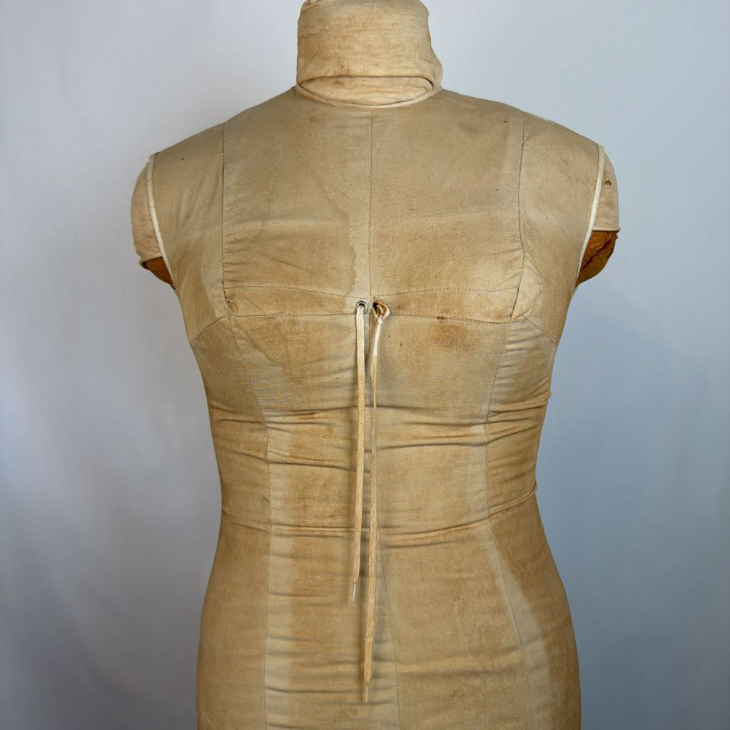 A beige/tan sleeveless mock-neck dress displayed on a fabric mannequin torso, showing the back view with a center tie and seam details against a plain light gray background.