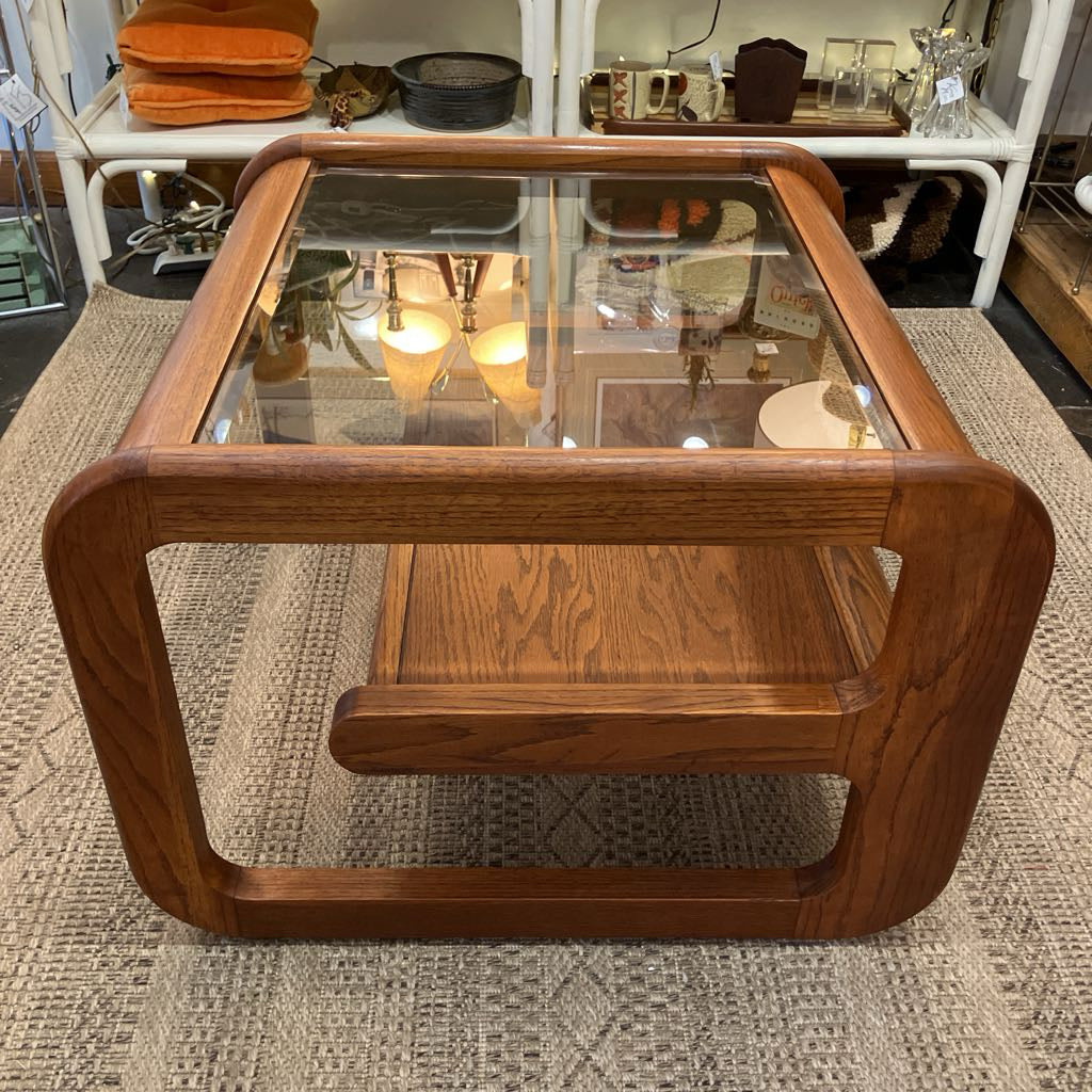 A mid-century modern wooden coffee table with a thick oak-look frame, a lower shelf, and a clear glass top, shown on a textured rug in a shop setting.
