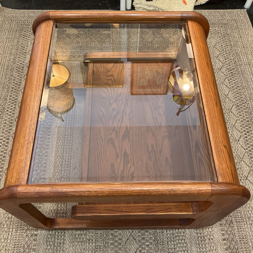 A square wooden coffee table with a thick glass top and rounded corners, resting on a textured beige rug.