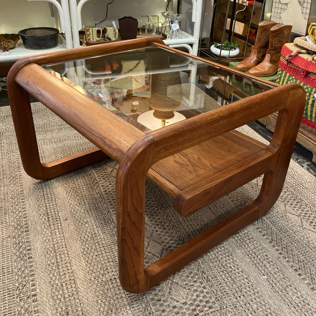 A mid-century modern wooden coffee table with a glass top, thick curved wooden arms, and a lower shelf, shown on a textured rug in a furniture store.