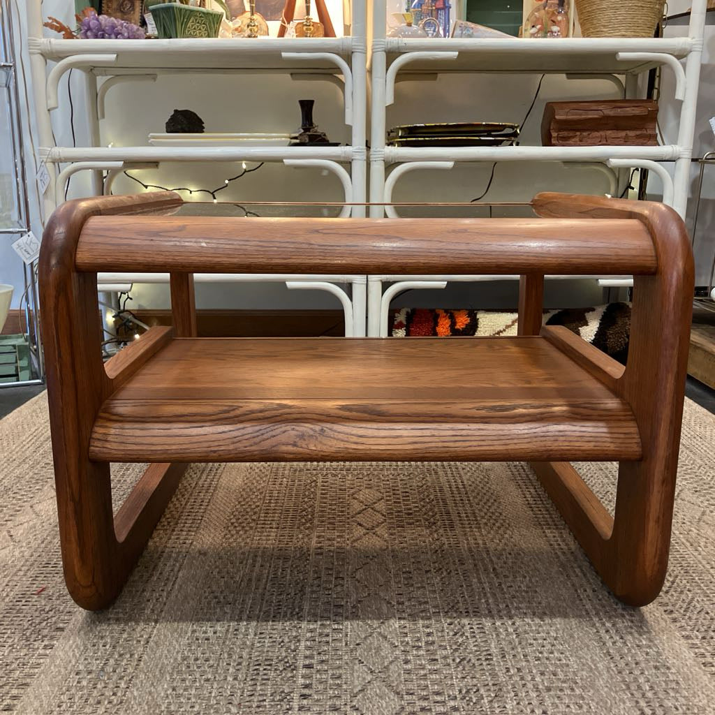 A small wooden folding table or shelf with a warm medium-brown finish, featuring a top rail, a middle shelf, and curved legs, set on a textured beige rug in front of a white shelving unit.