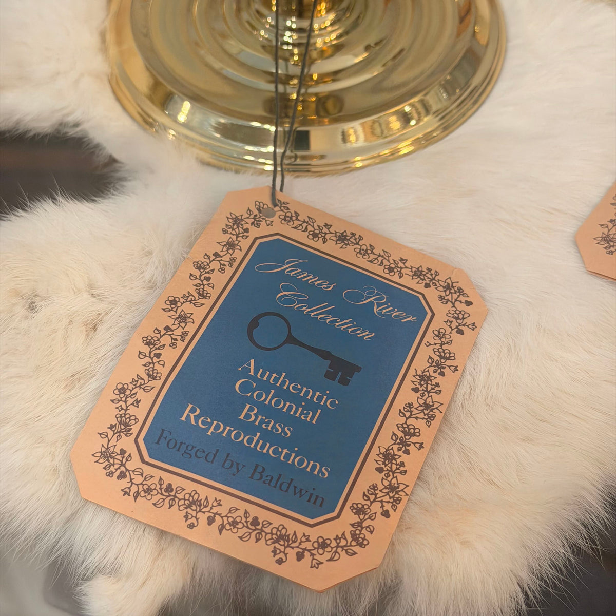 A hanging paper tag for the James River Collection Authentic Colonial Brass Reproductions, styled on a white faux fur surface with a gold brass candle holder base in the background.