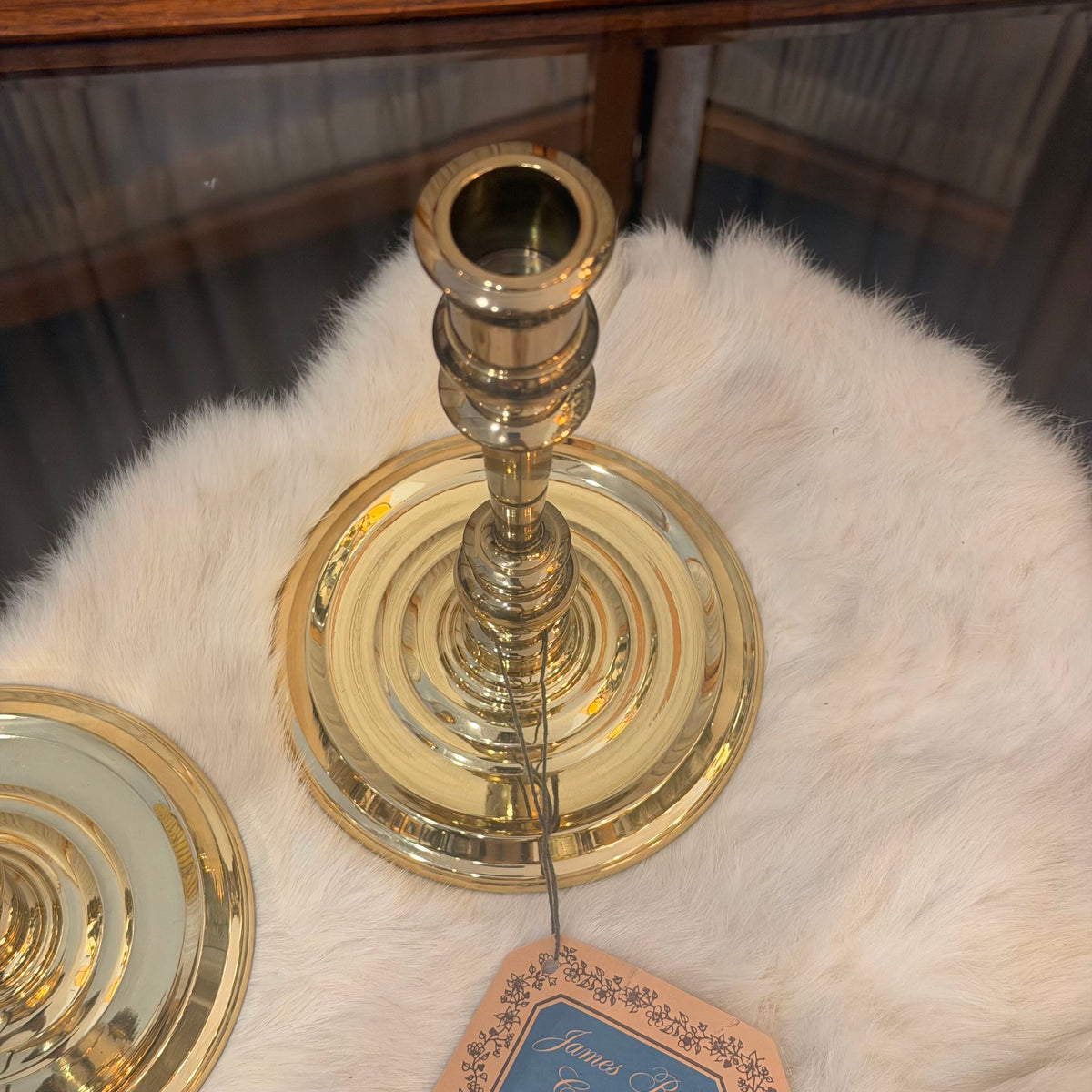 A polished brass candlestick holder with a circular base and tiered stem, displayed on a white faux fur surface with part of another matching piece visible at the lower left.