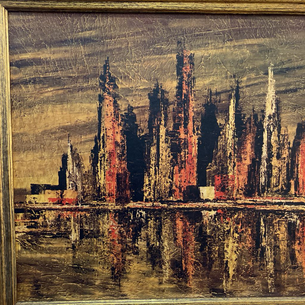 A framed abstract cityscape painting featuring tall spires and reflections in dark brown, black, red, and gold tones, with a visible wooden frame.