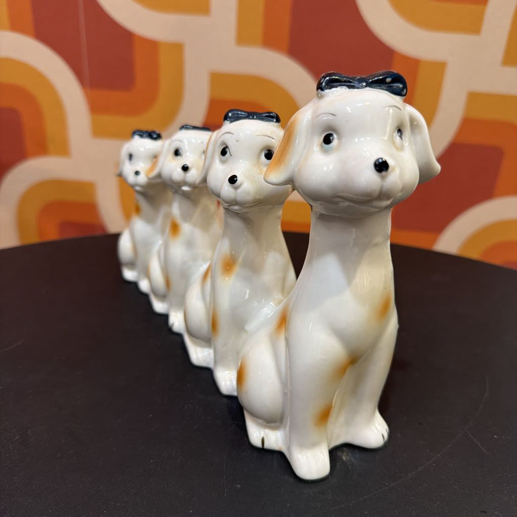 A row of five glossy ceramic dog figurines in white with orange patches and black bows, displayed on a dark surface against a retro orange and cream patterned background.