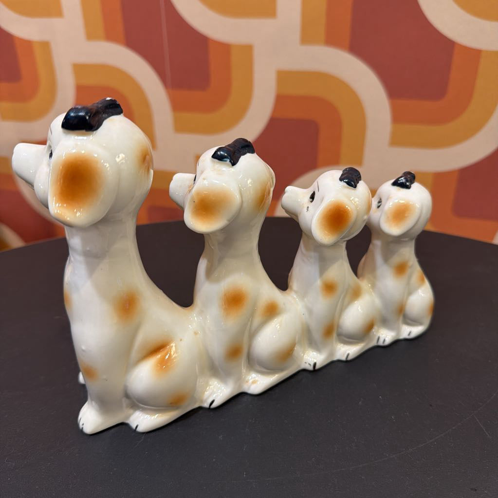 A set of four ceramic dalmatian dog figurines in a row, decorated with white glaze, orange spots, and black ears and noses, displayed on a dark surface against a retro orange and cream patterned background.