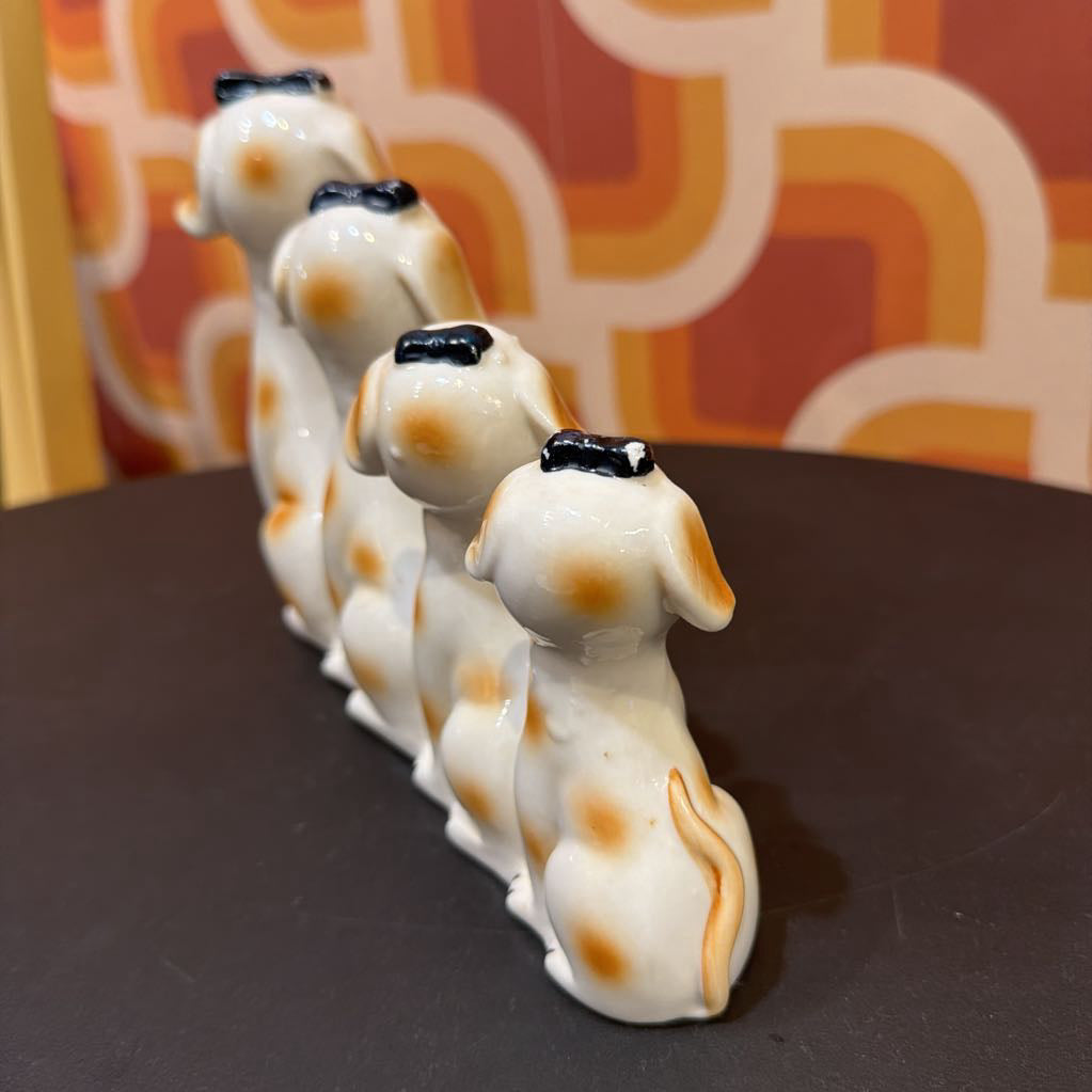 A set of five glossy ceramic dog figurines lined up in a row, featuring a white base with orange spots and black caps, displayed on a dark surface against a patterned orange and yellow background.