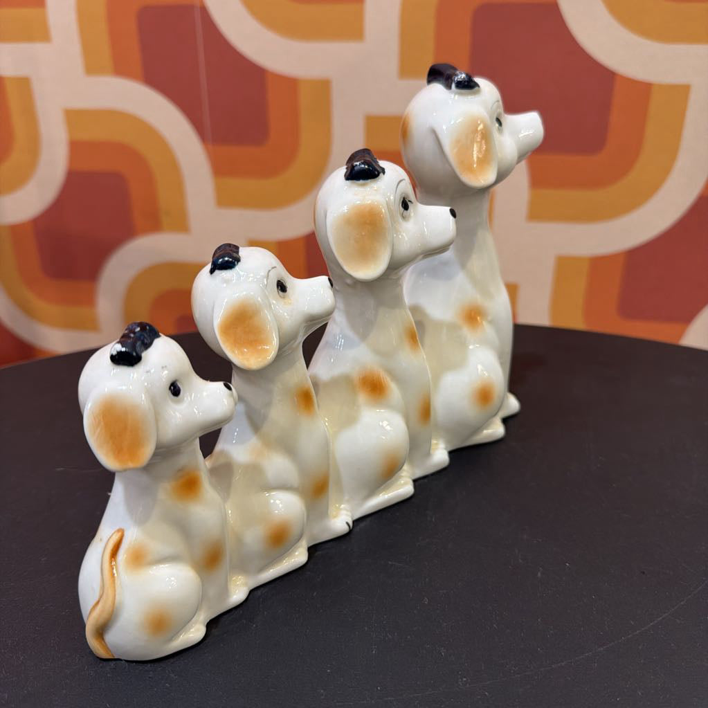 Four ceramic dalmatian dog figurines arranged in a row on a dark tabletop, with a retro orange and beige patterned background.