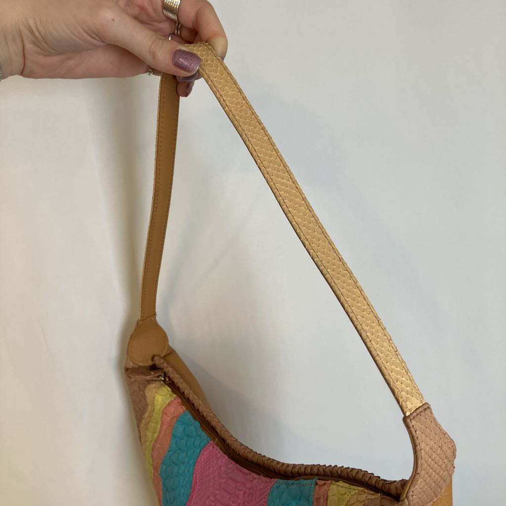A hand holding the shoulder strap of a crescent-shaped shoulder bag with a multicolor patchwork quilted body and tan leather strap, set against a plain off-white background.