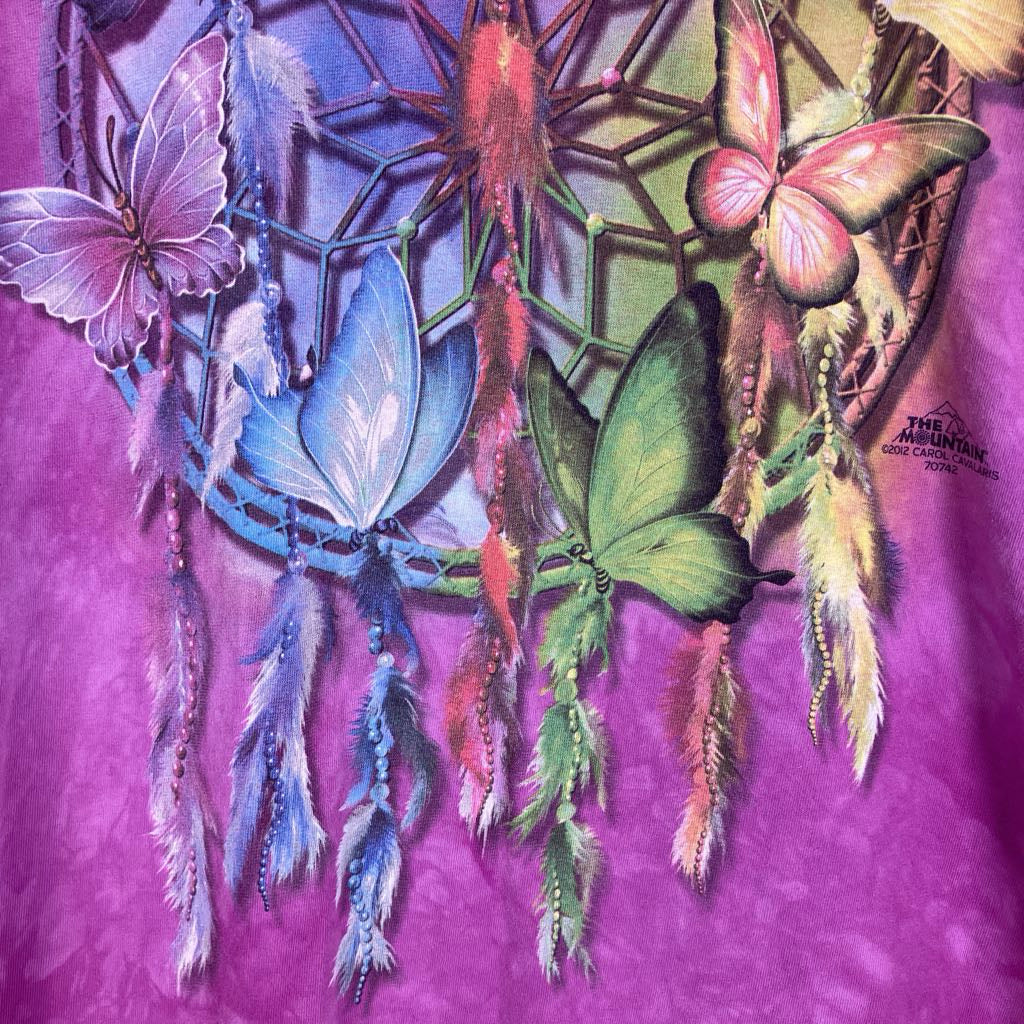 A close-up of a purple garment featuring a colorful dreamcatcher and butterfly graphic. The design includes blue, green, pink, and orange butterflies with hanging feathers, and a small printed logo reading 'The Amazing Grace Apparel' on the right side.