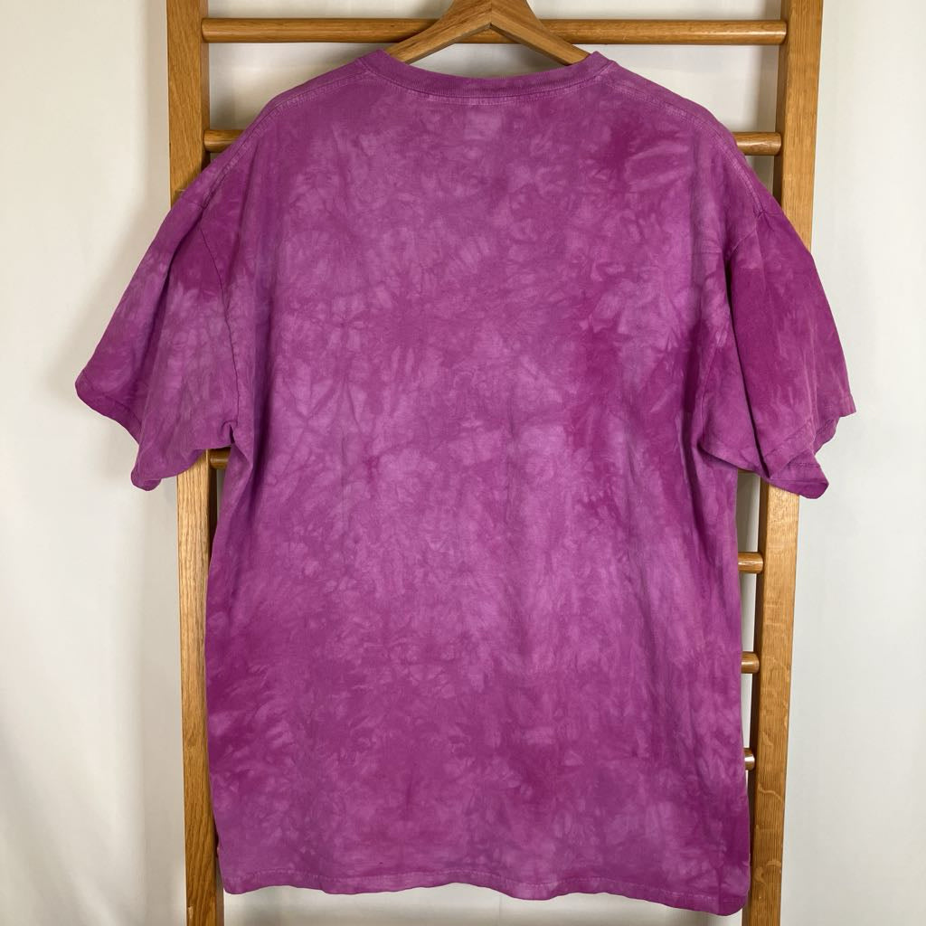 A purple tie-dye short-sleeve t-shirt hanging on a wooden hanger from a wooden clothes rack against a plain off-white wall.