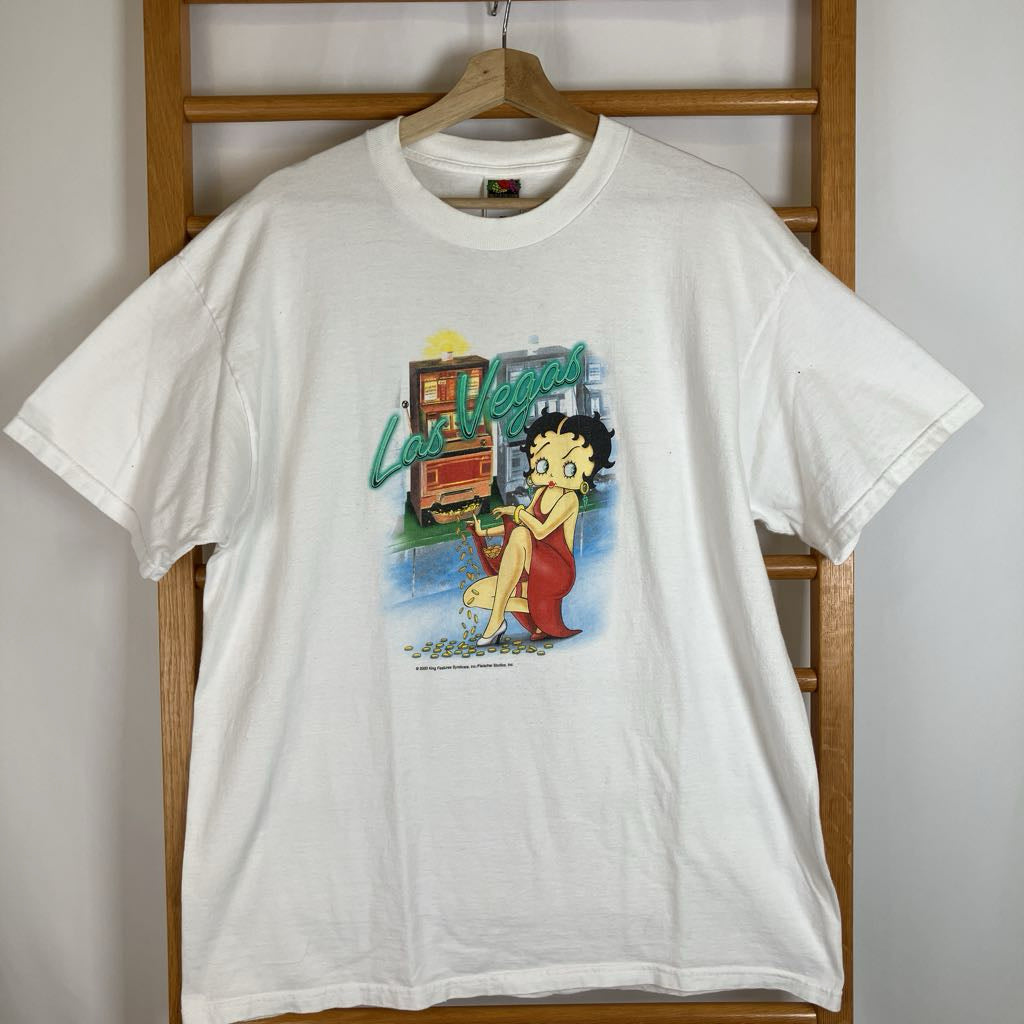 A white vintage graphic t-shirt featuring Betty Boop and the 'Las Vegas' sign, hanging on a wooden hanger against a wooden chair back.
