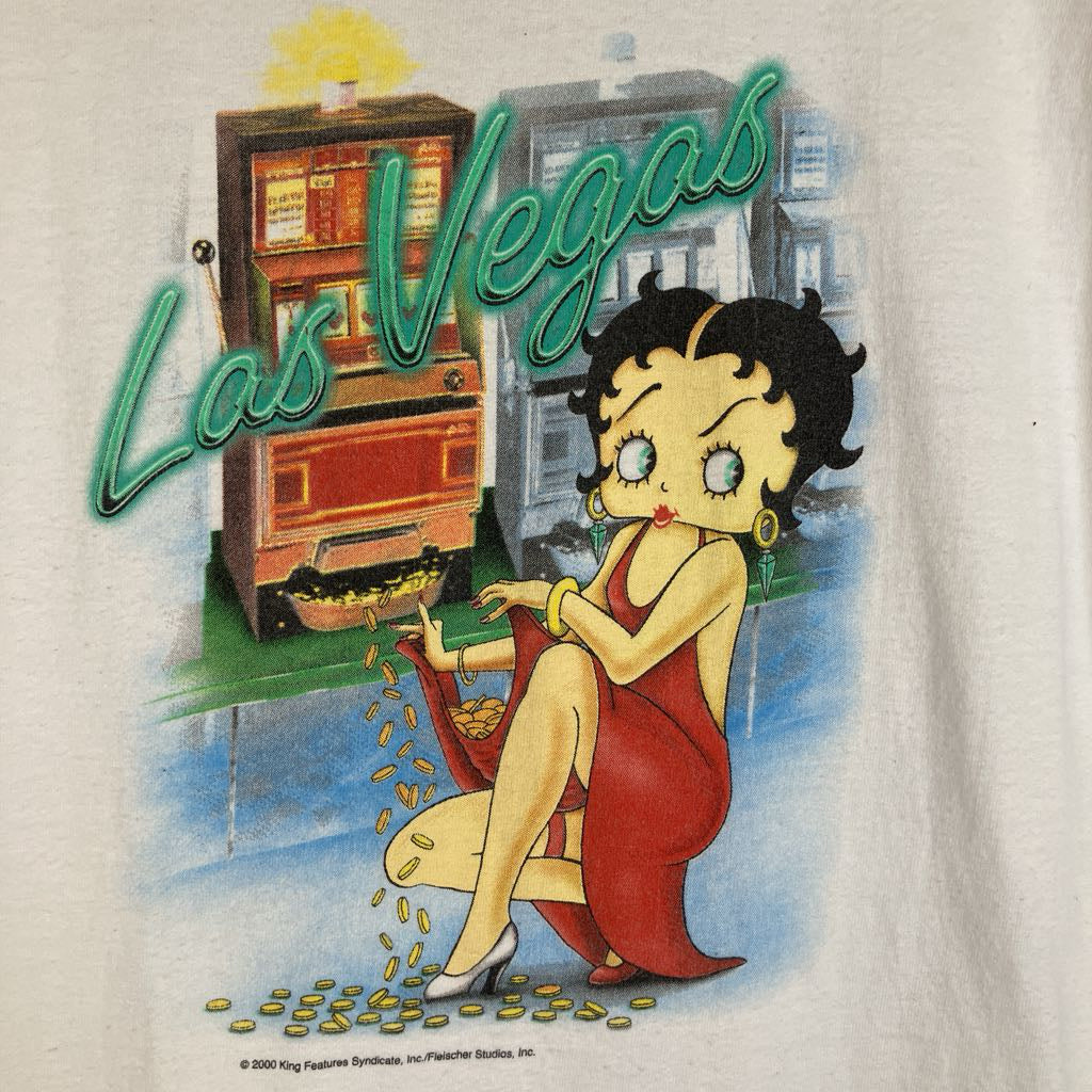 A close-up of a vintage-style graphic print on a light fabric background, featuring the words 'Las Vegas' in large teal script above an illustrated cartoon woman in a red dress sitting beside a slot machine with gold coins.
