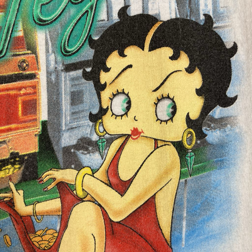 A close-up of a graphic print featuring the cartoon character Betty Boop wearing a red dress, seated in front of a neon sign and a building. The print appears on a light-colored fabric surface.