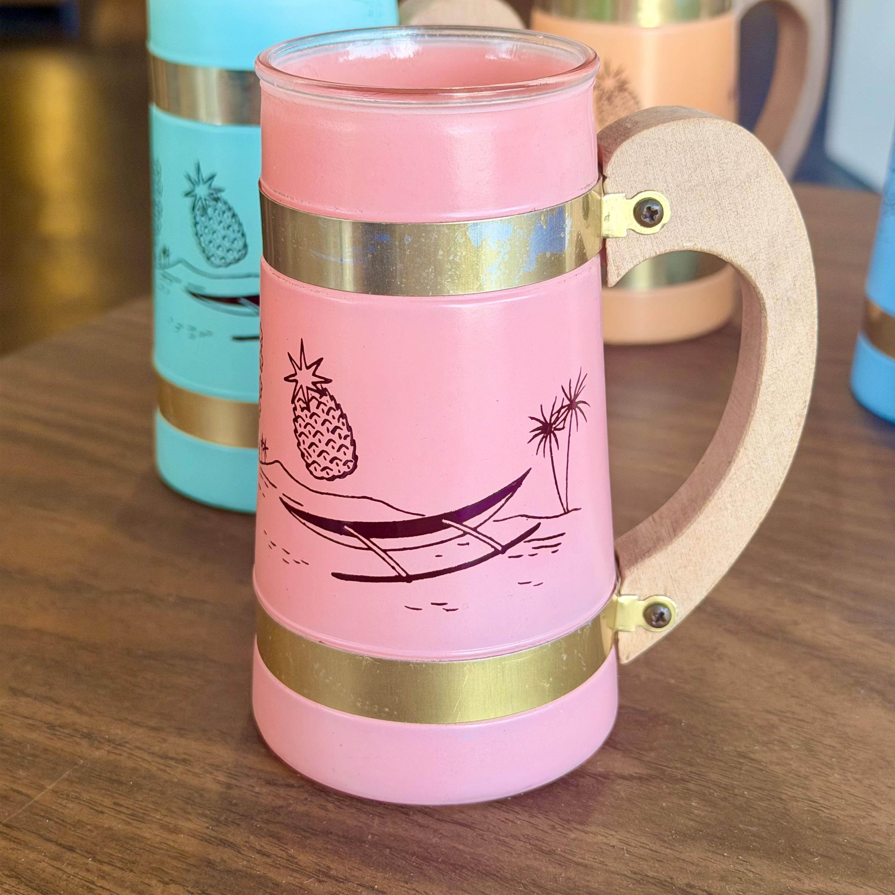 A pink ceramic mug with a light wood handle and gold metal bands, decorated with a black line art pineapple and beach scene. The mug is shown on a wooden table with other mugs blurred in the background.