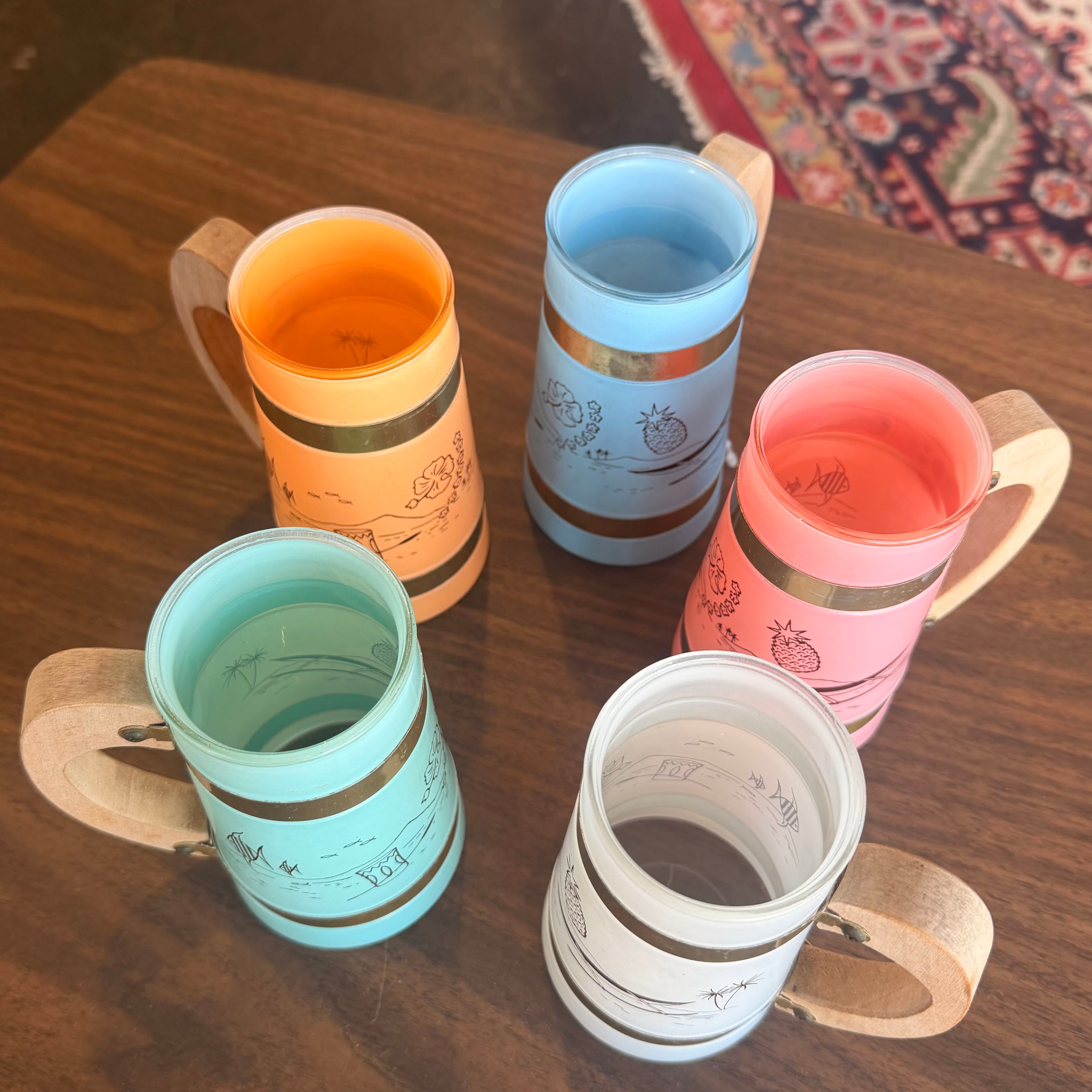 Four handled cups in pastel colors arranged on a wooden table. The cups are orange, light blue, pink, and white, each decorated with simple line drawings and metallic-looking bands near the rim.