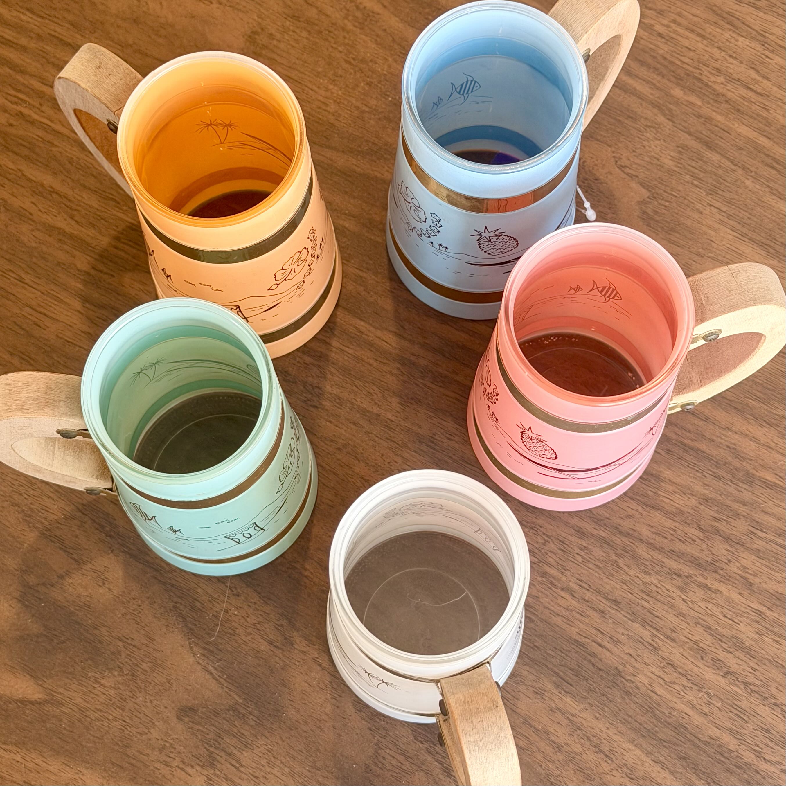 Five ceramic tumblers with wooden handles arranged on a wooden tabletop. The tumblers have pastel-colored interiors and exteriors with gold bands and simple line drawings.