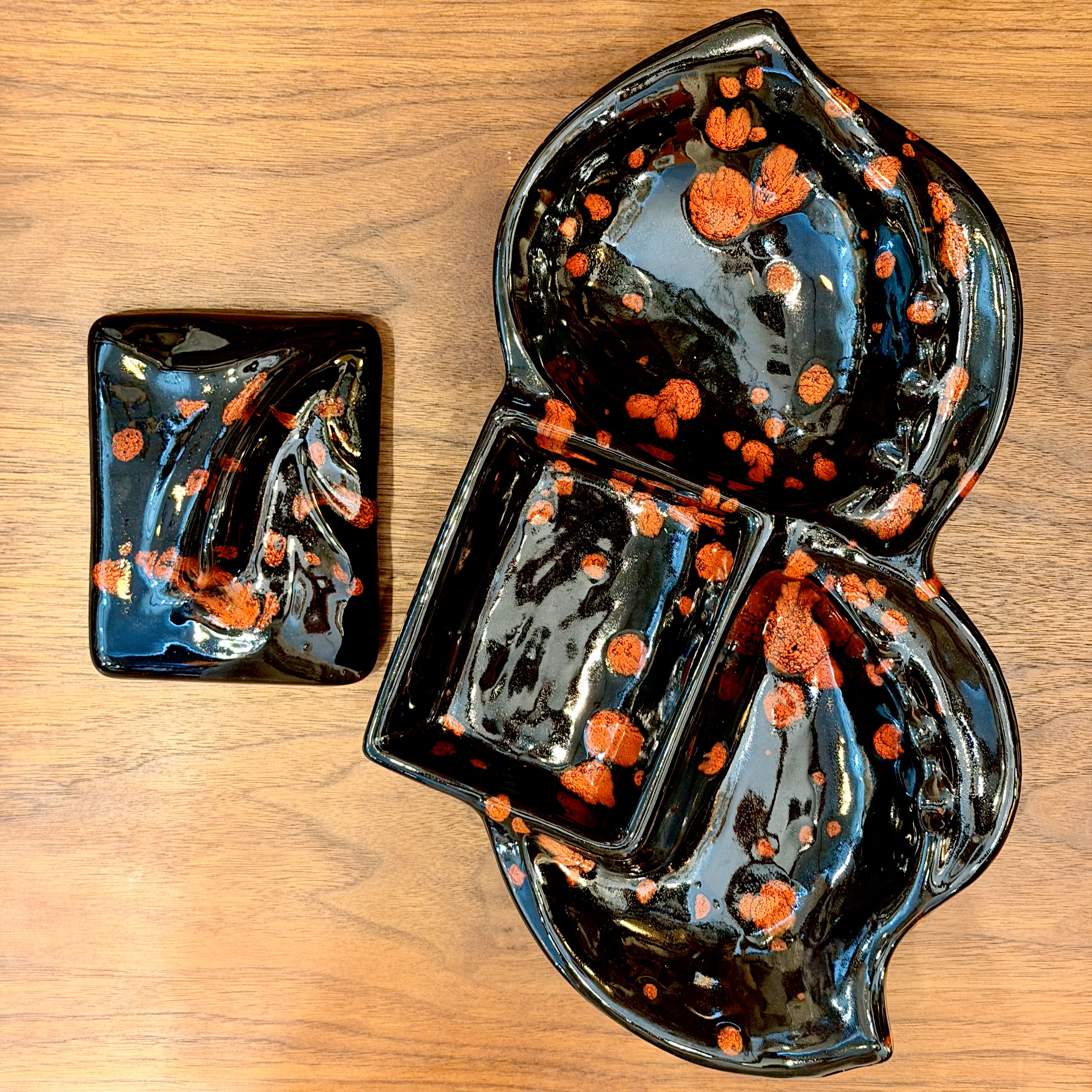 A glossy black ceramic serving piece with orange speckled accents, shown on a wooden tabletop. The set includes a large divided serving platter and a smaller matching dish.