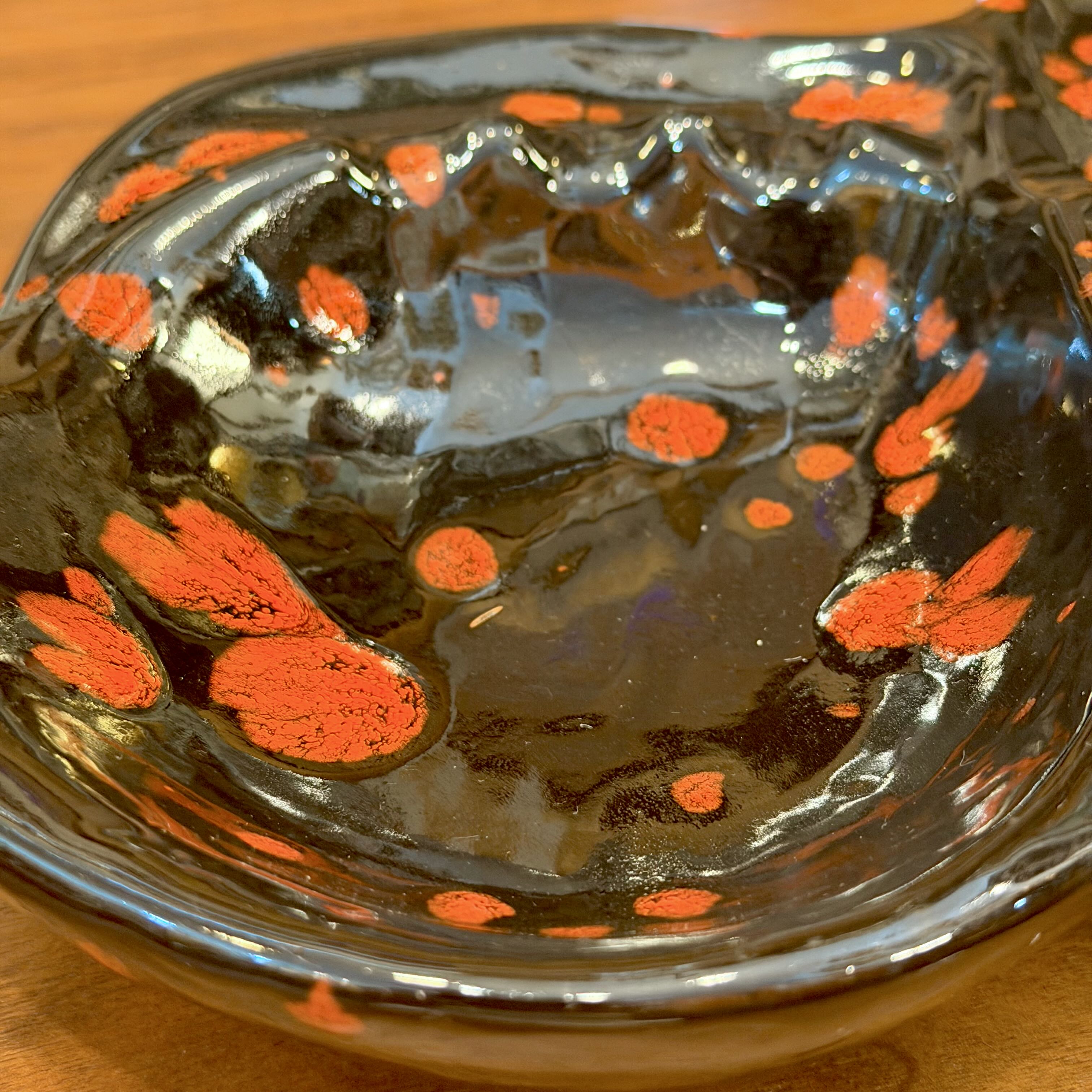 A close-up of a glossy black ceramic bowl with orange abstract painted spots and splatters, resting on a wooden surface.
