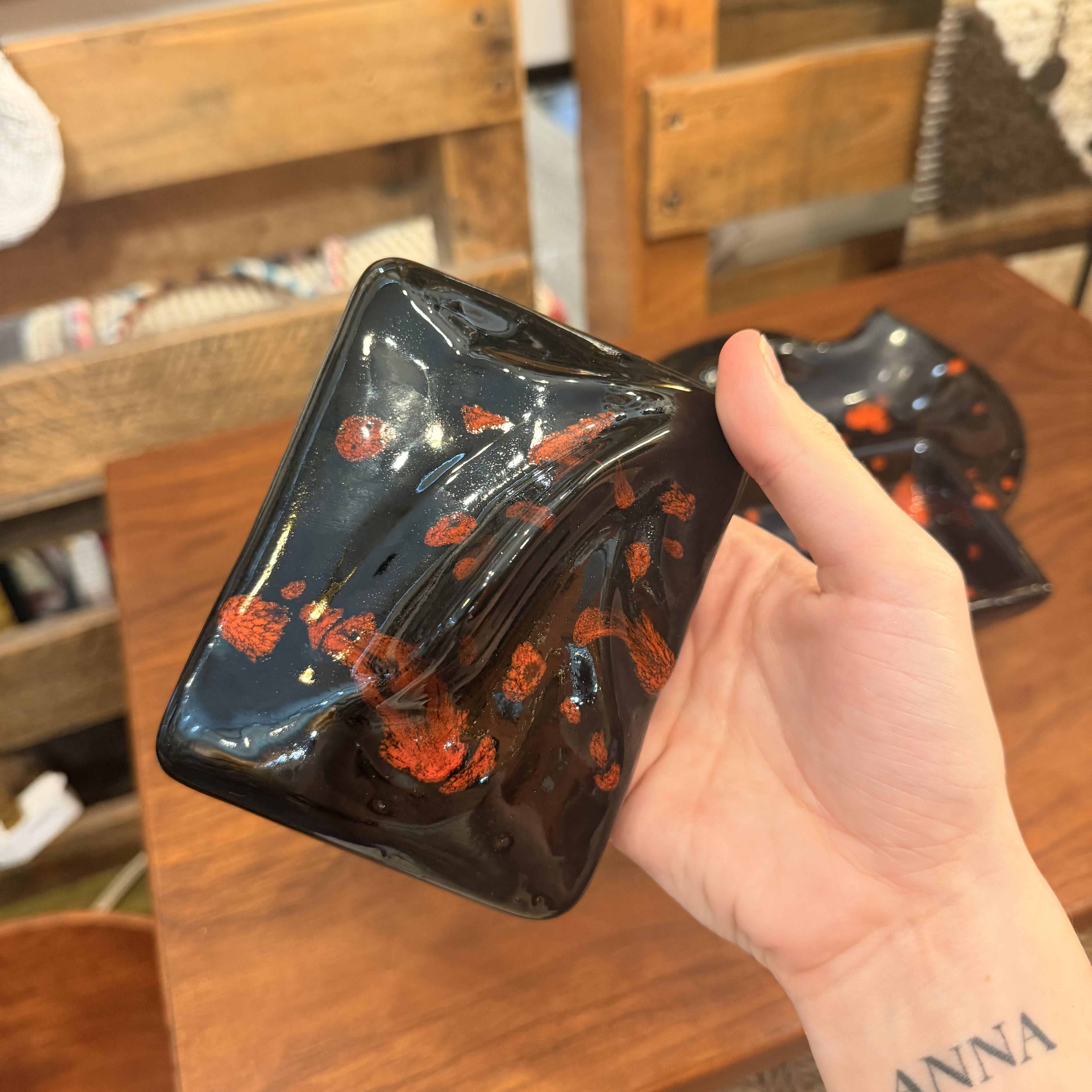 A hand holds a glossy black glass dish with orange-red speckled patterns, with another matching dish blurred in the background on a wooden table.