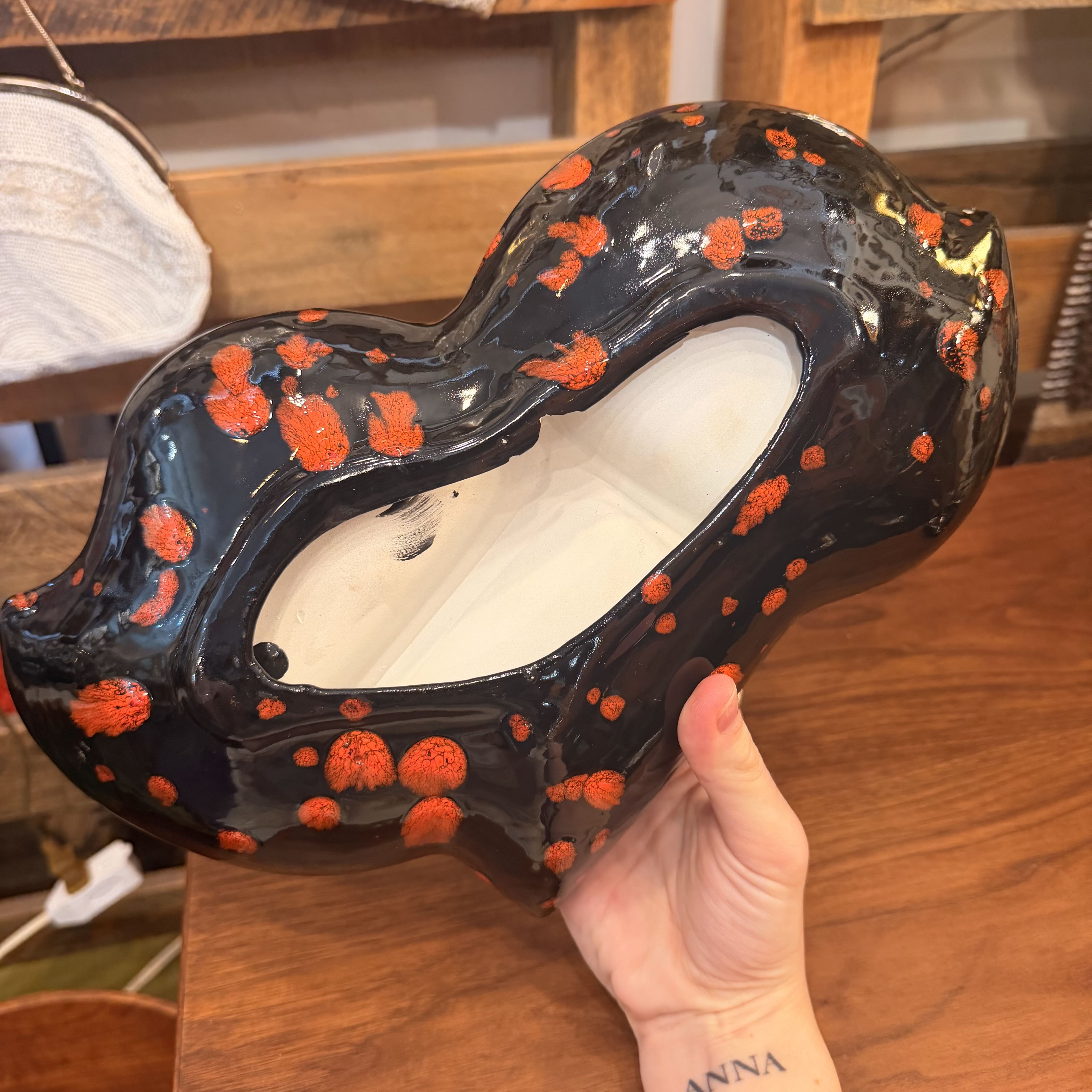 A hand holds a glossy black ceramic planter shaped like a stylized animal head, decorated with orange-red speckled spots, against a wooden background.