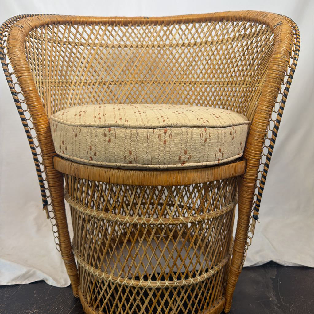 A vintage-style woven rattan armchair with a tall curved backrest and a cylindrical base, featuring a beige cushion with a small geometric pattern on the seat.