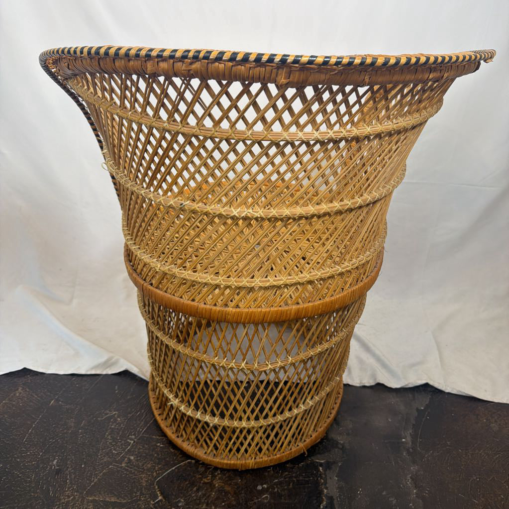 A natural woven rattan basket with a flared rim and diamond-pattern weave, standing on a dark surface against a white fabric backdrop.